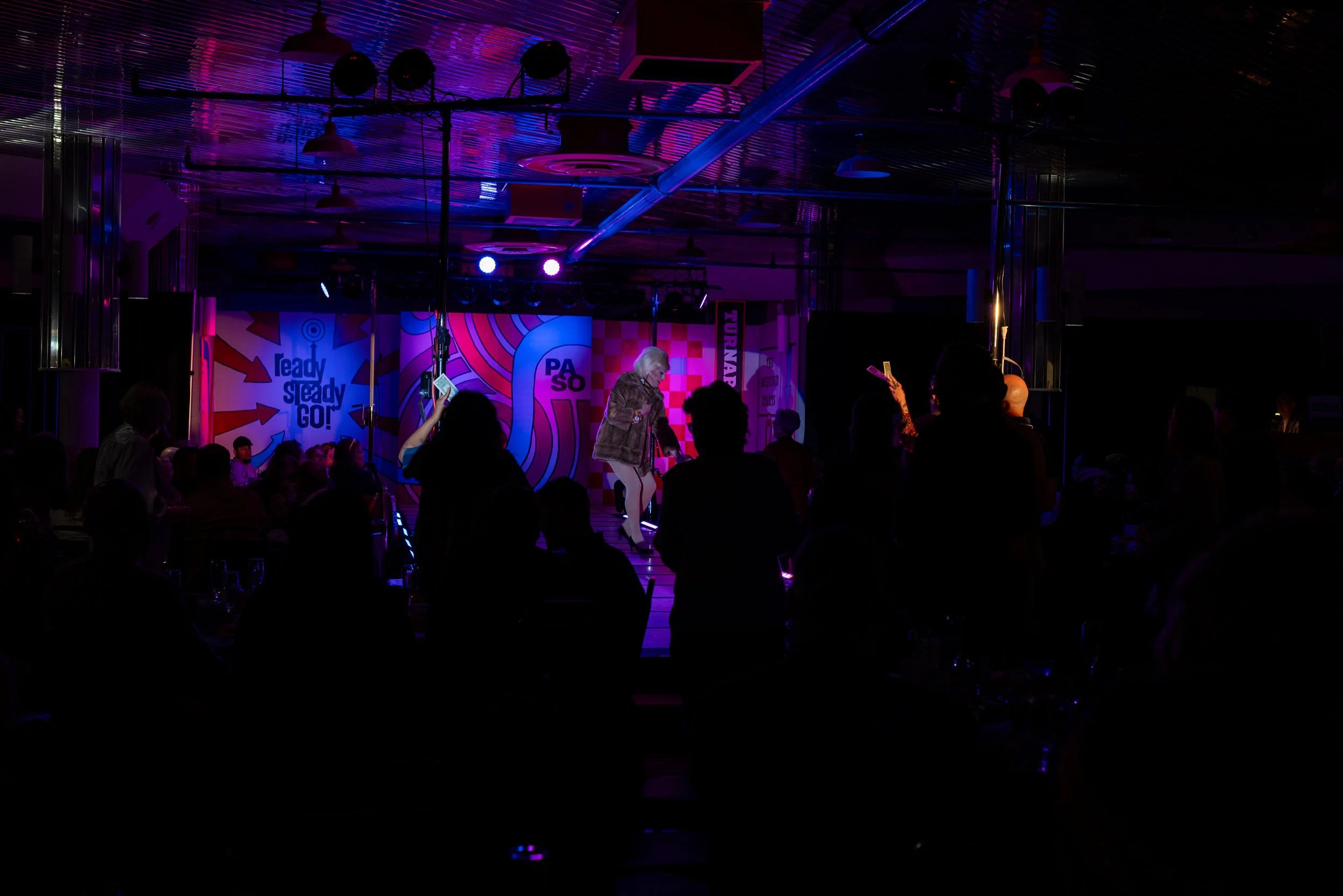 A lively indoor performance with a woman singing on stage, surrounded by an audience in a dimly lit room with colorful neon lights and music-themed decorations.