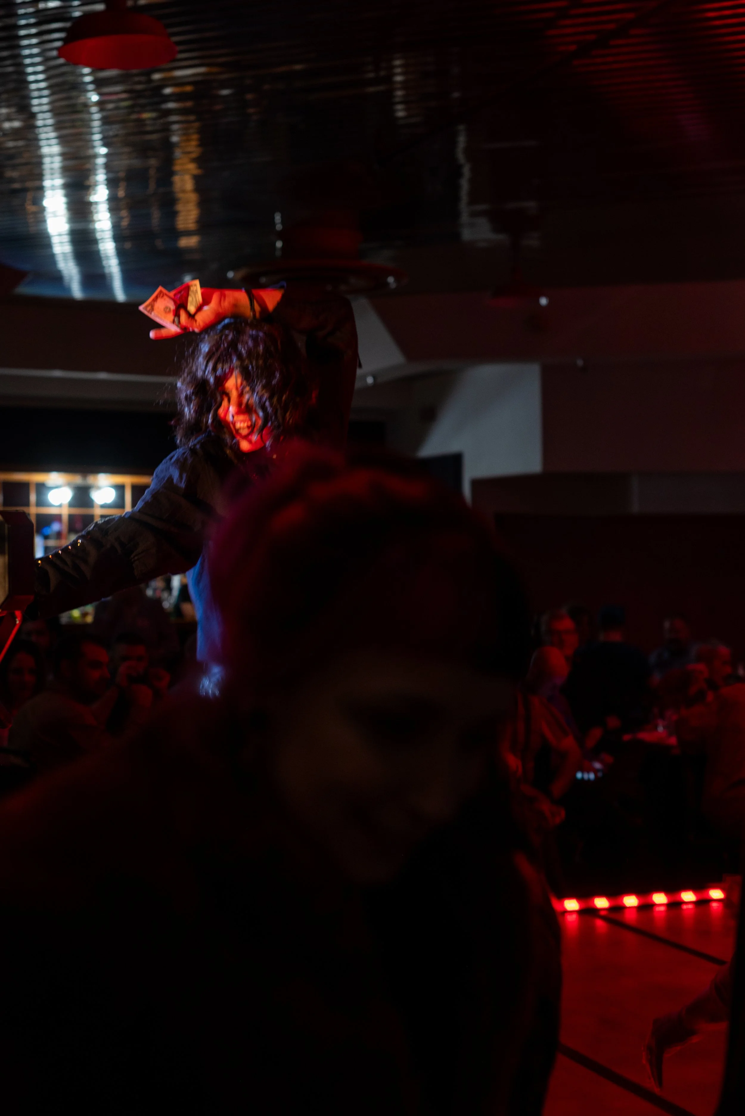 A woman with curly hair and painted face dancing and holding money in a dark, red-lit venue with an audience watching.