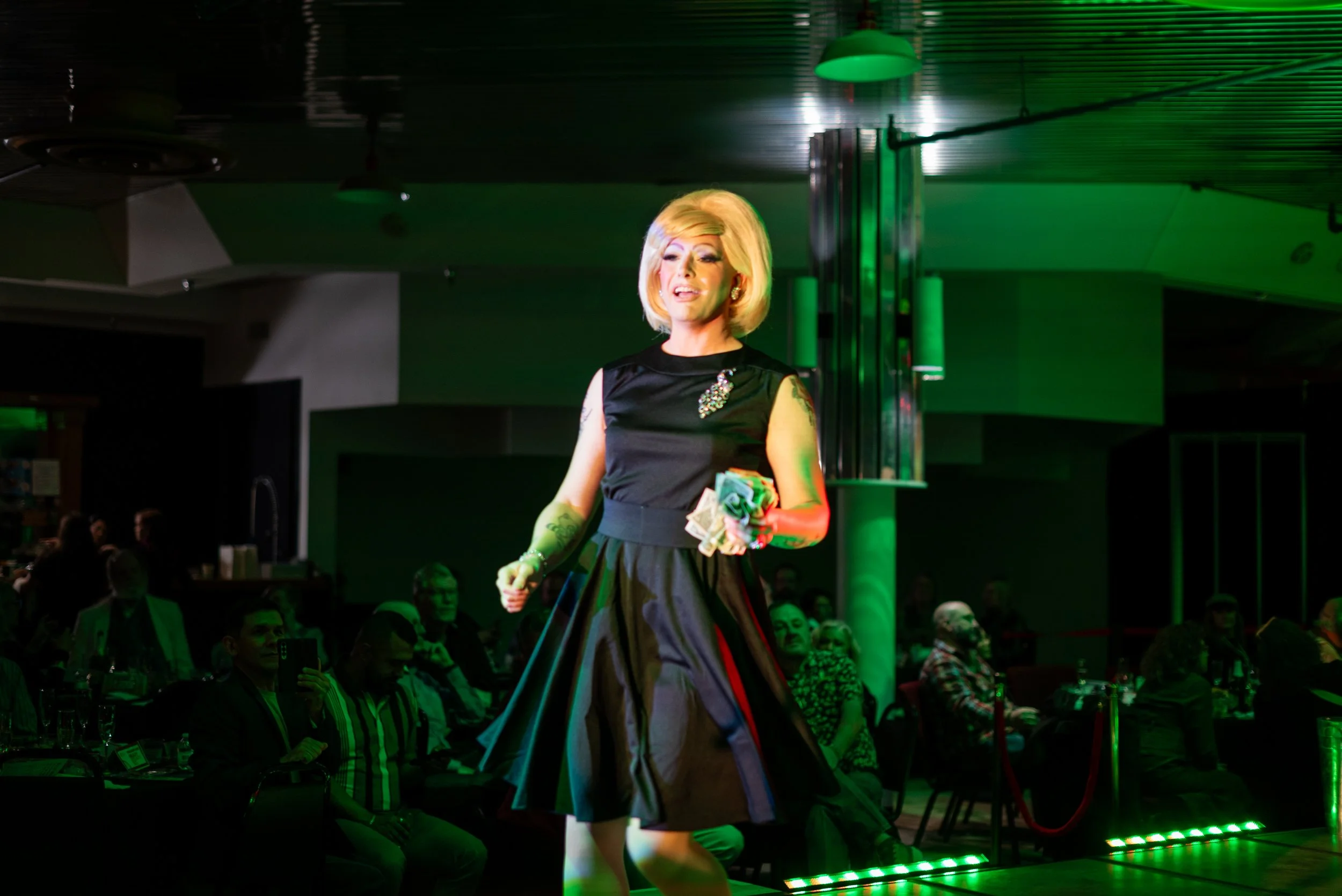 A person with blonde hair and vintage style clothing, wearing a black dress with a decorative brooch, standing on stage in front of an audience inside a venue with green lighting.