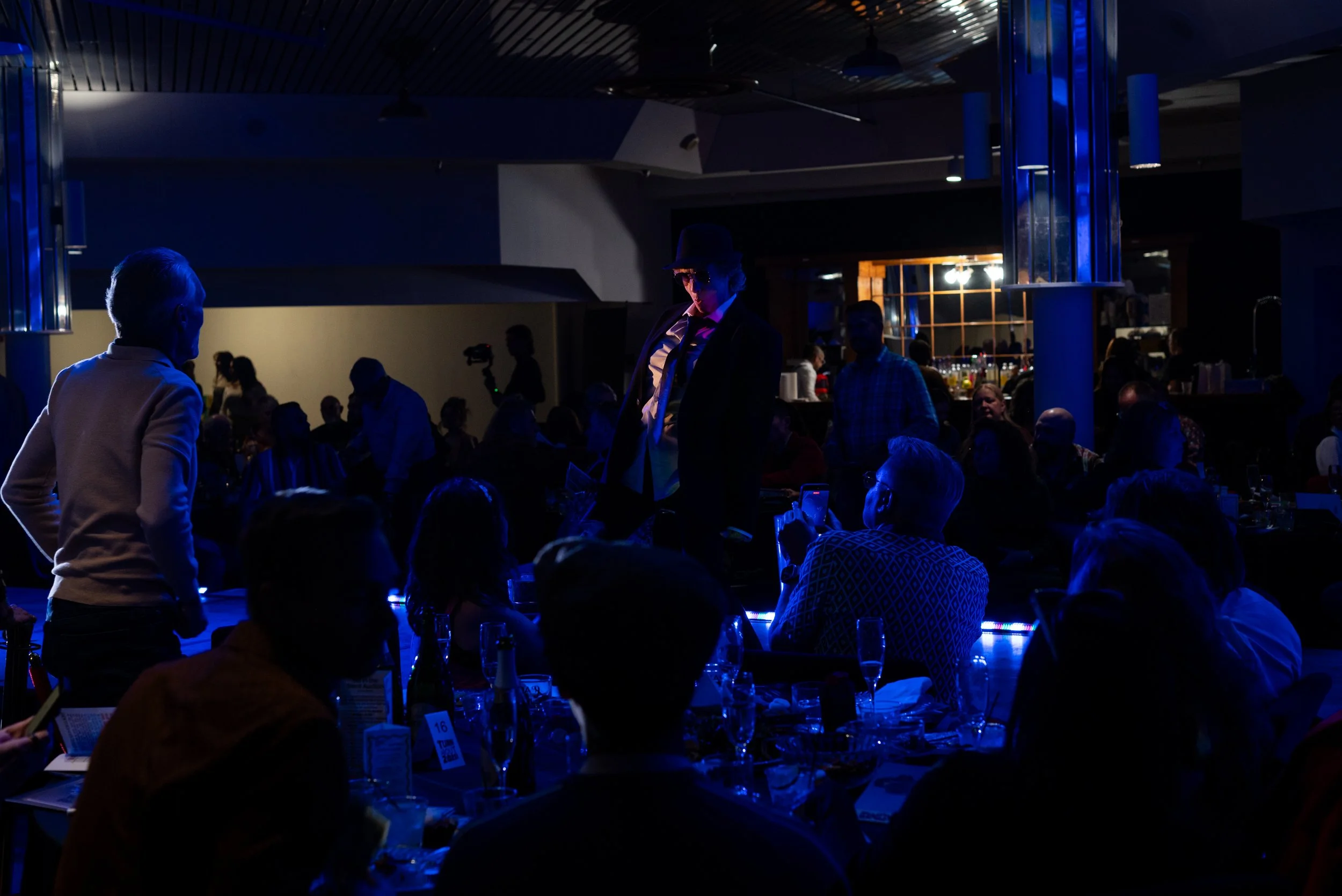 People gathered in a dimly lit indoor venue, with a person in a suit and hat standing on a stage or platform. Rest of the scene includes individuals sitting at tables, some using phones, and a bar area in the background.