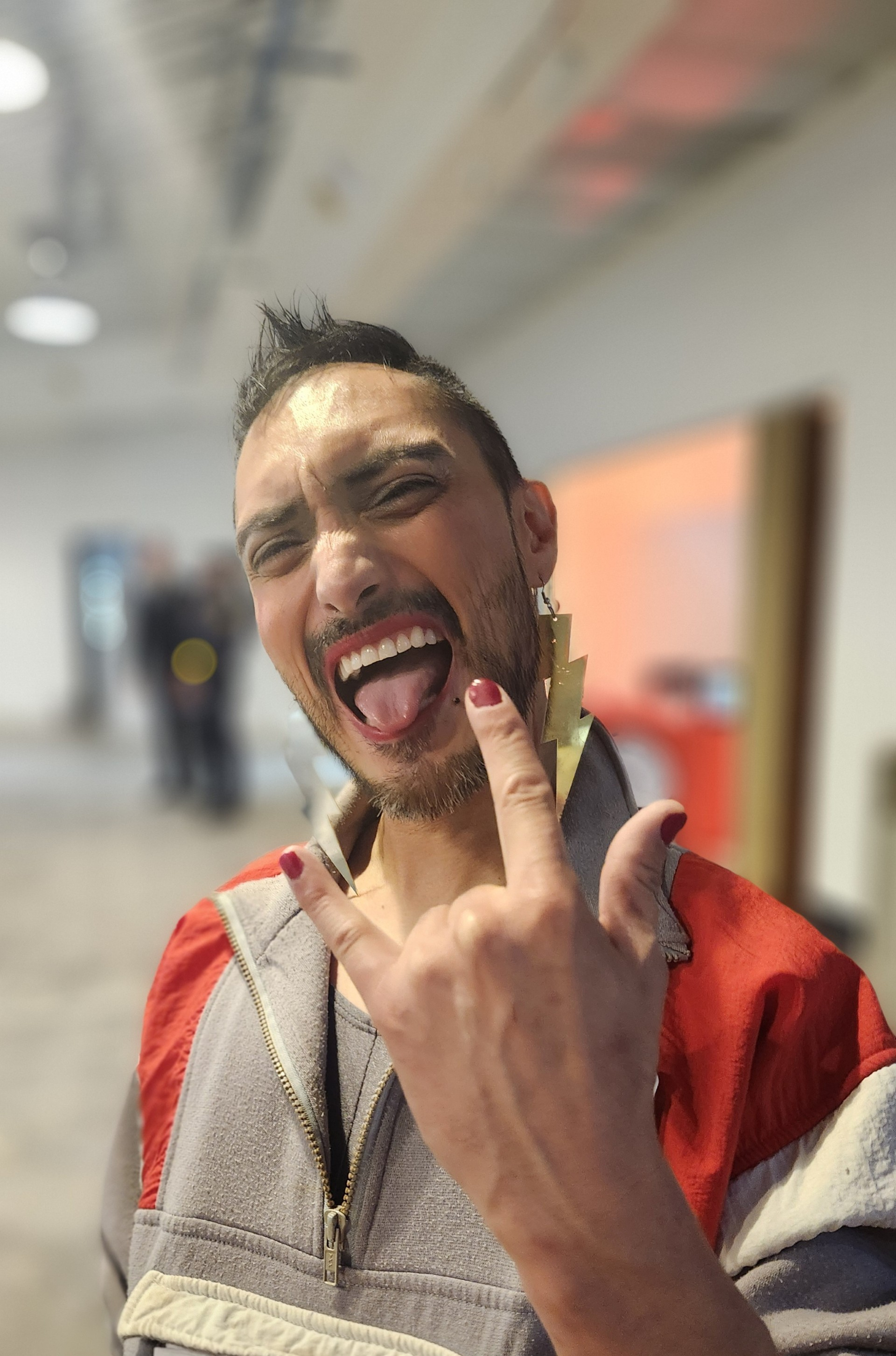A man with short dark hair and a beard making a face, sticking out his tongue and squinting, while pointing with his finger to his large gold lightning bolt earring. He is wearing a zip-up jacket with color blocking, including gray, red, and cream. The background is blurry with some indistinct figures and objects.