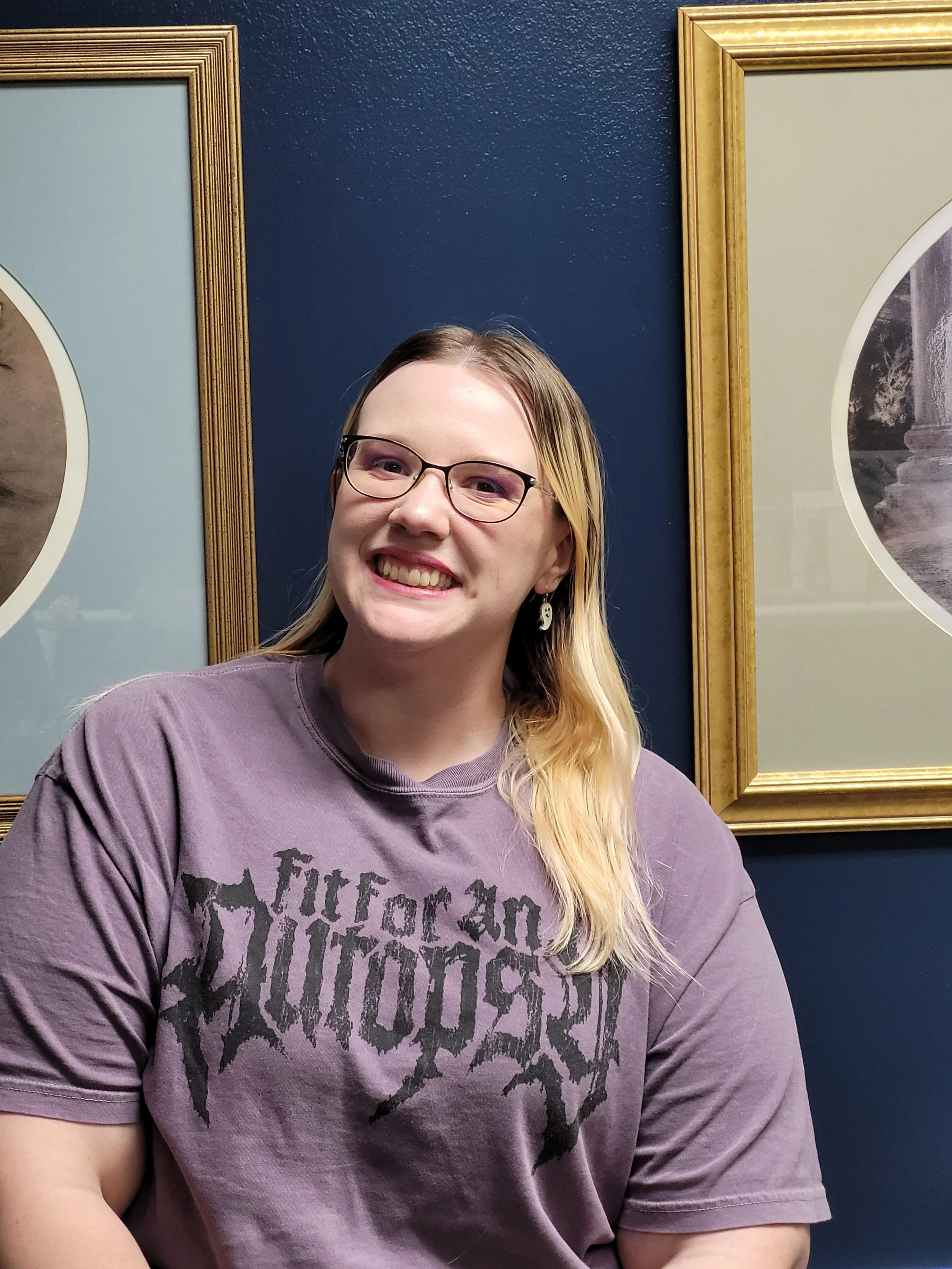 A woman with glasses and long blonde hair smiling, wearing a purple graphic t-shirt, seated in front of a blue wall with framed artwork.