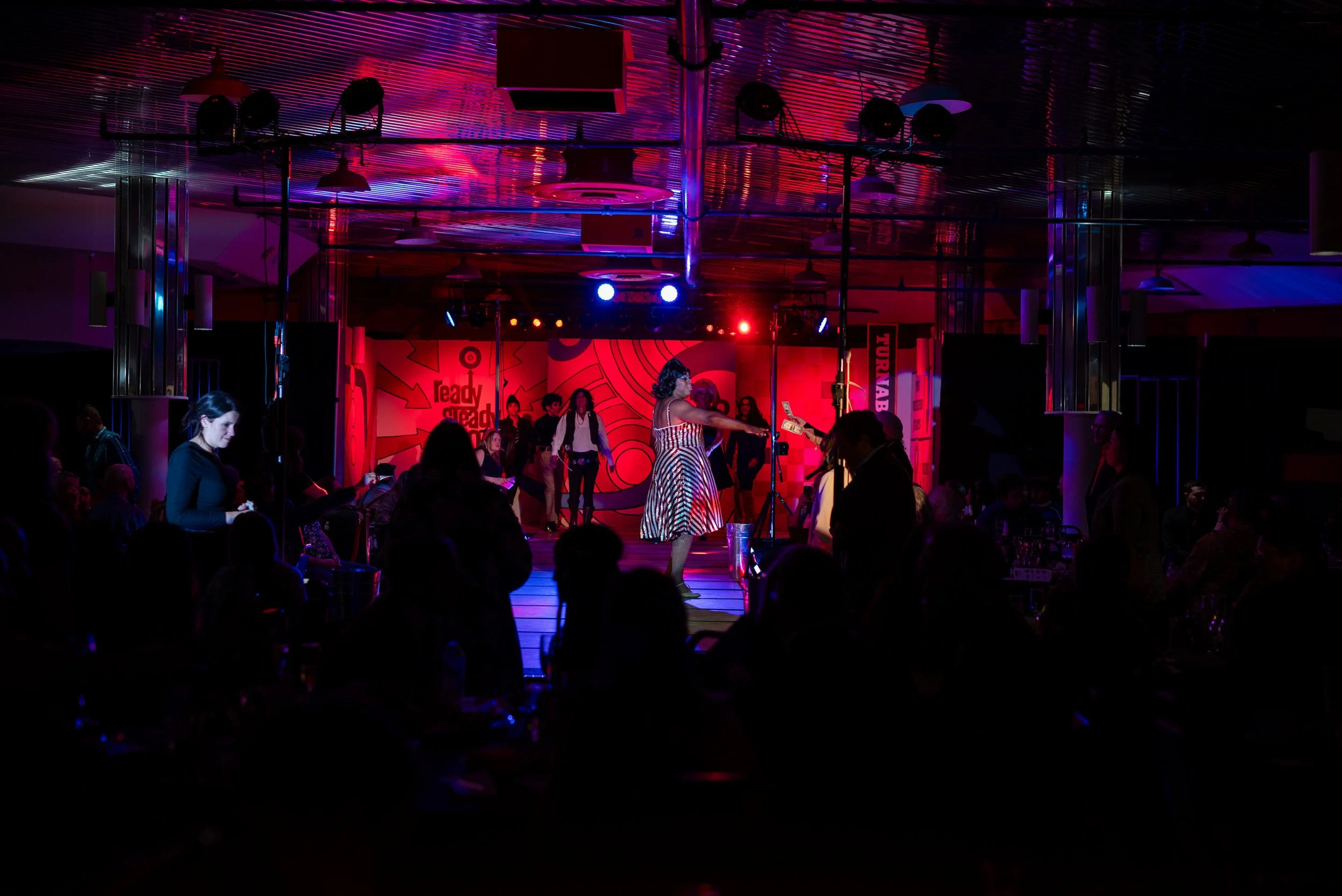 A woman performing on stage in a colorful striped dress at a lively event with an audience, stage lighting, and a decorated backdrop.