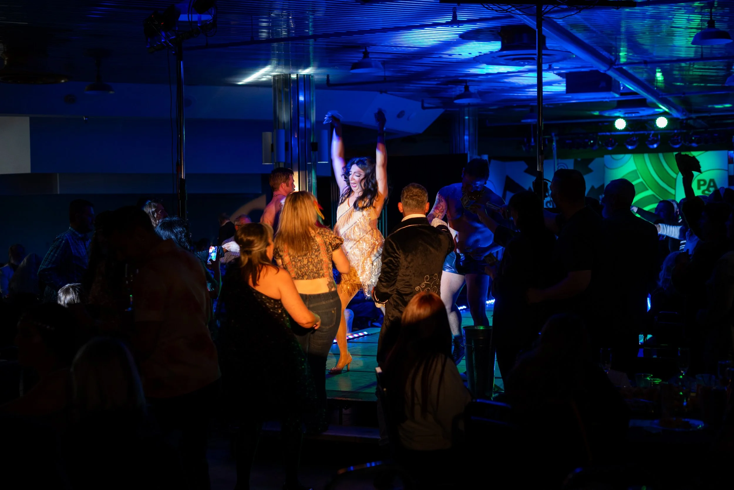 A woman dancing on a stage with raised arms, surrounded by a crowd in a dimly lit nightclub or bar with colorful lighting.