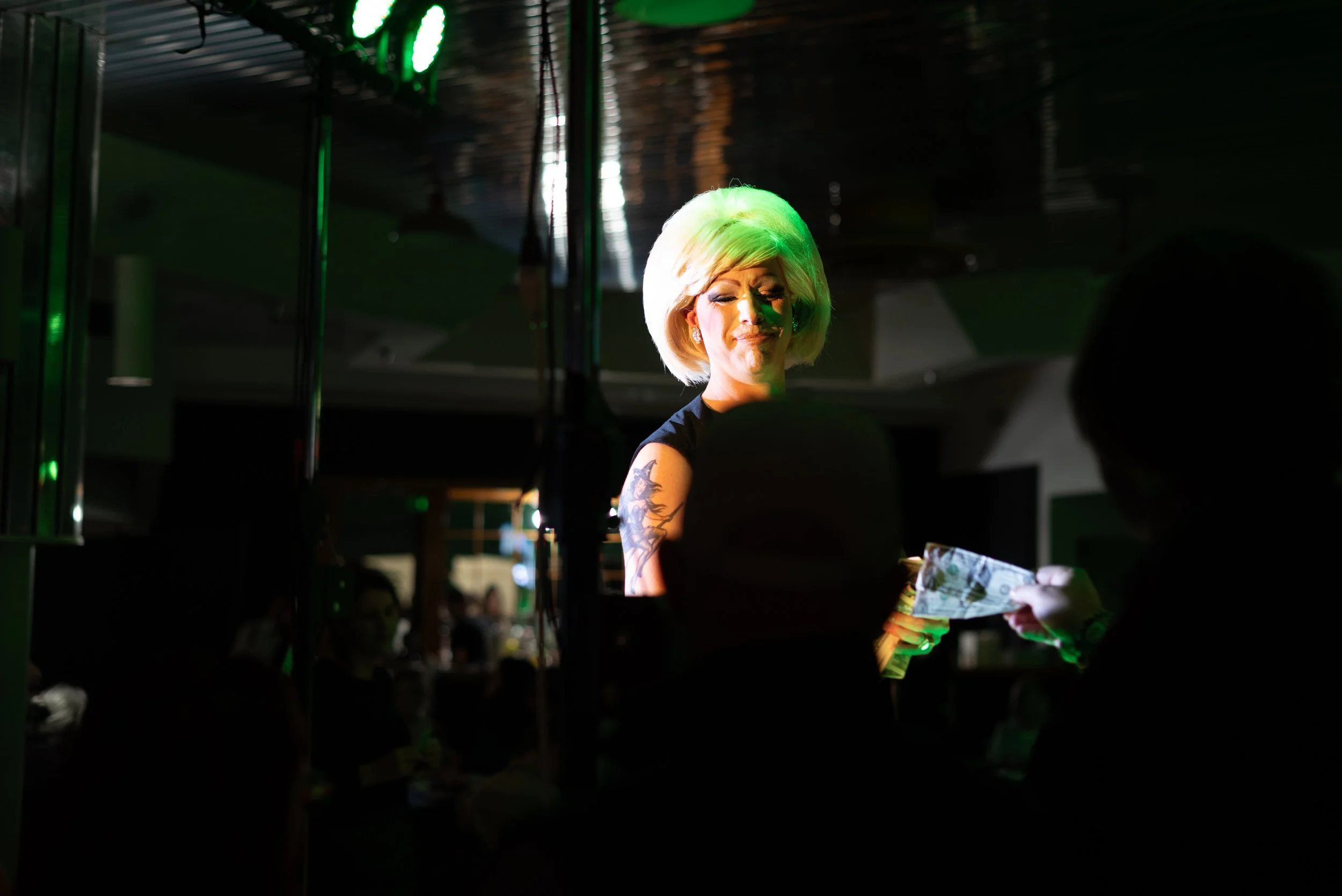 A woman with blonde hair standing in a dimly lit venue, receiving cash from someone. She has a tattoo on her arm and is illuminated by green lighting.