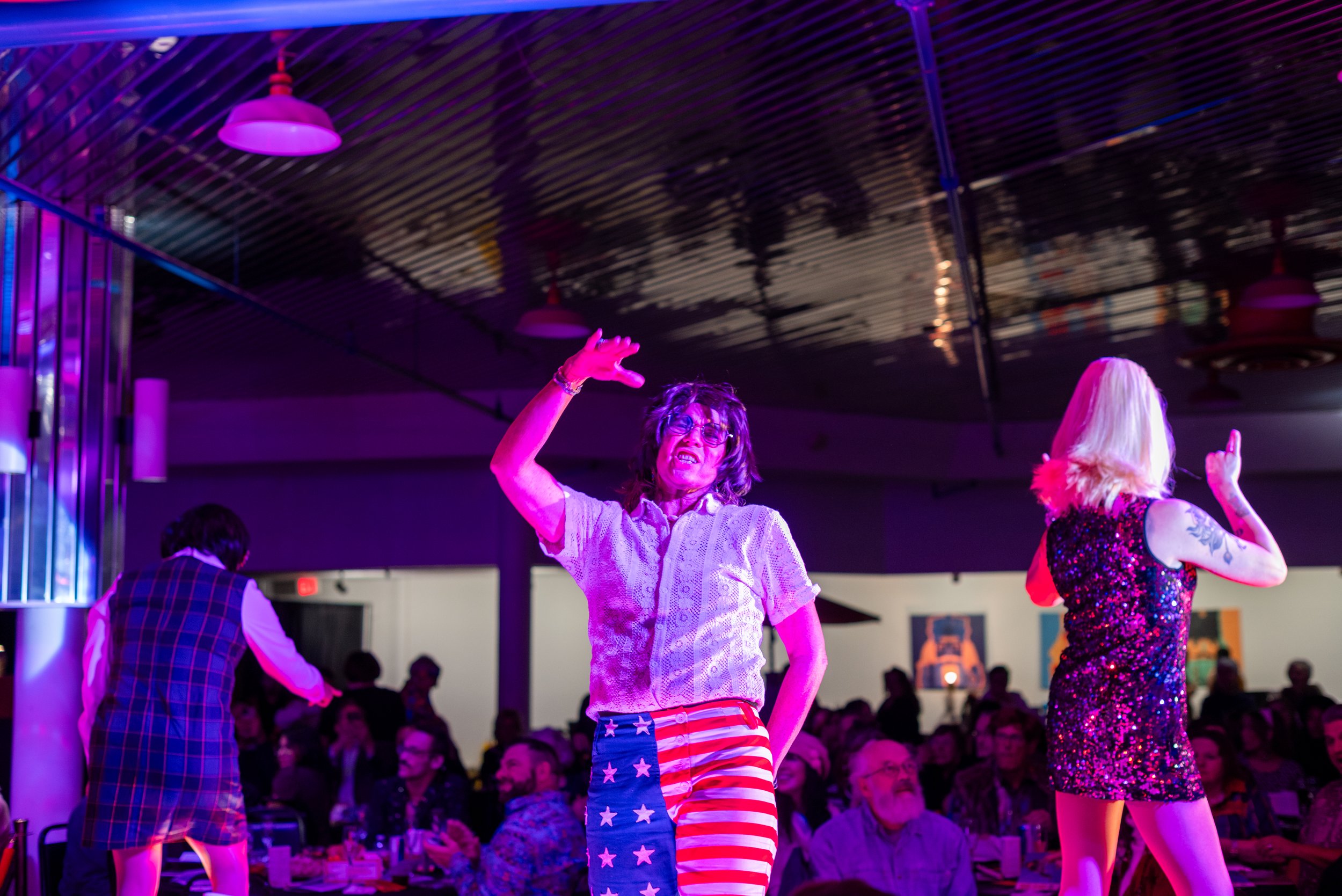 People performing on stage at a lively event with audience in the background, colorful lighting, and vibrant atmosphere.
