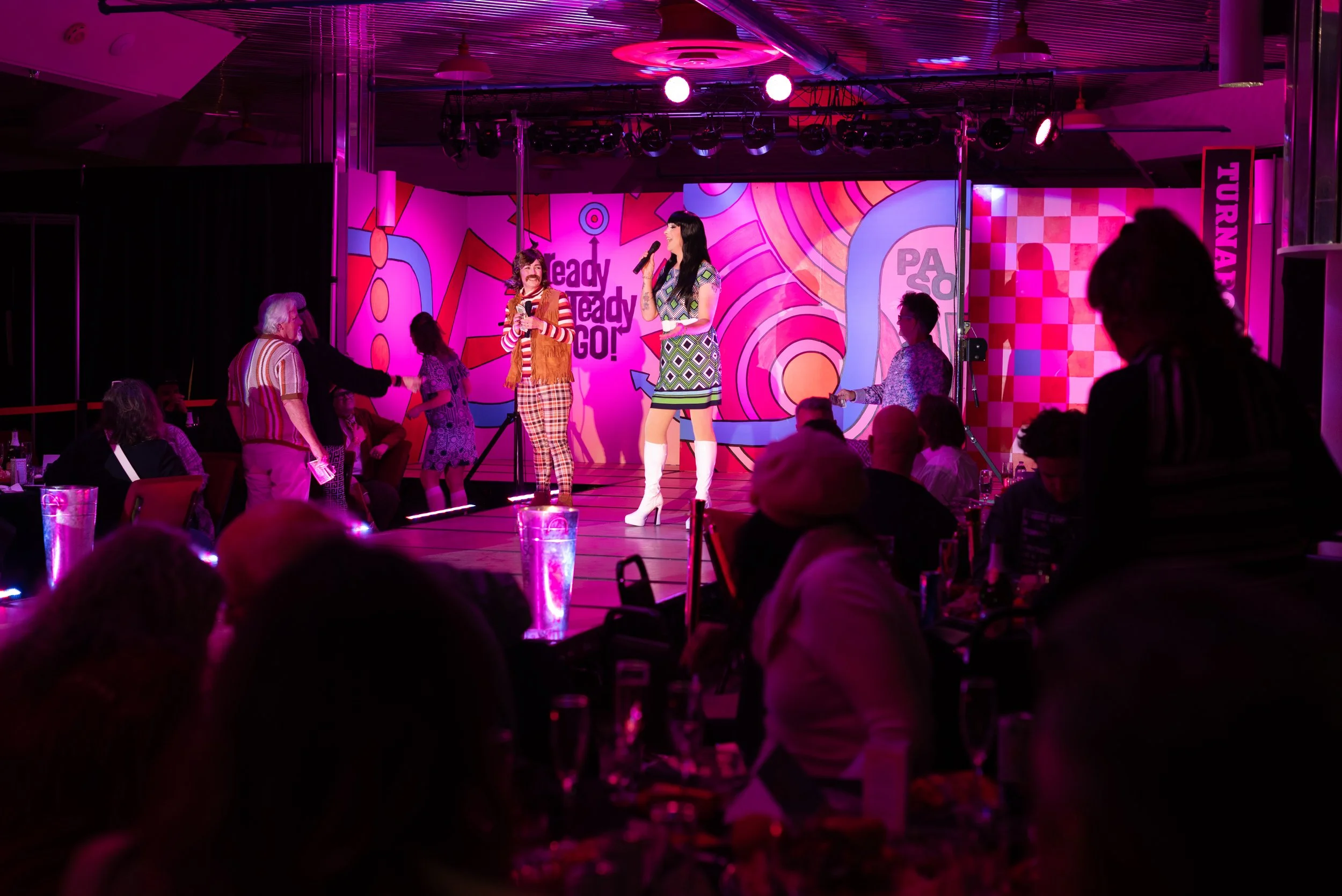 People performing and socializing at a karaoke event on a brightly lit stage with vibrant, colorful background graphics.
