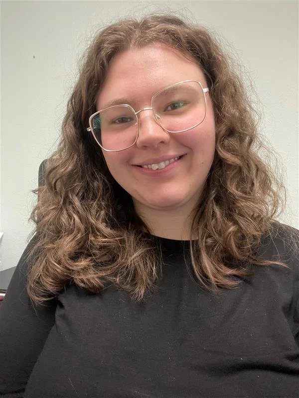 A woman with curly brown hair wearing glasses and a black shirt, smiling at the camera, in front of a beige wall.