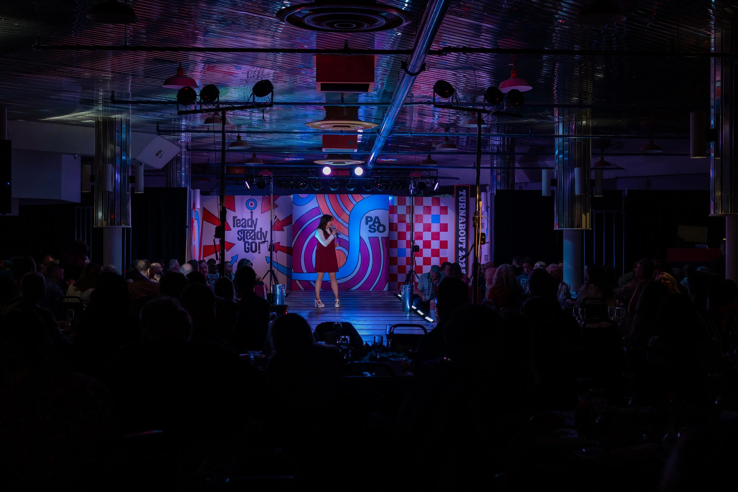 A woman performing on stage at an event with a colorful backdrop that says 'ready steady GO!' and 'PASO.' The audience is seated in front of the stage in a dimly lit venue with black curtains and overhead lighting fixtures.
