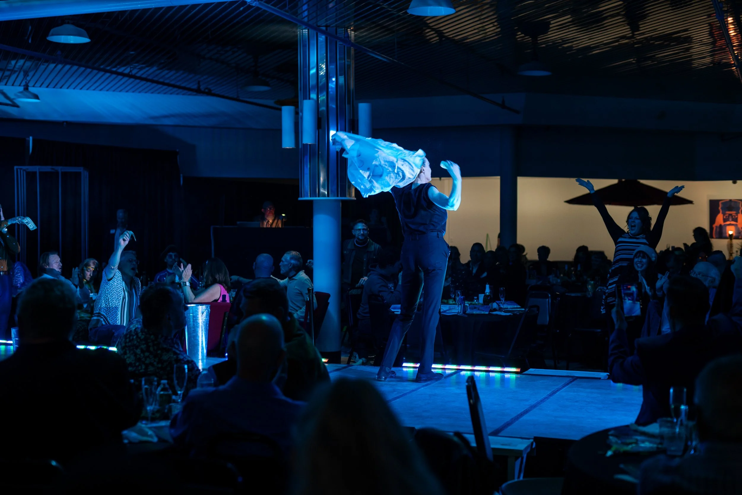 Performer on stage with a piece of fabric in a dark, blue-lit venue, surrounded by an audience watching and clapping, with some raising their hands.