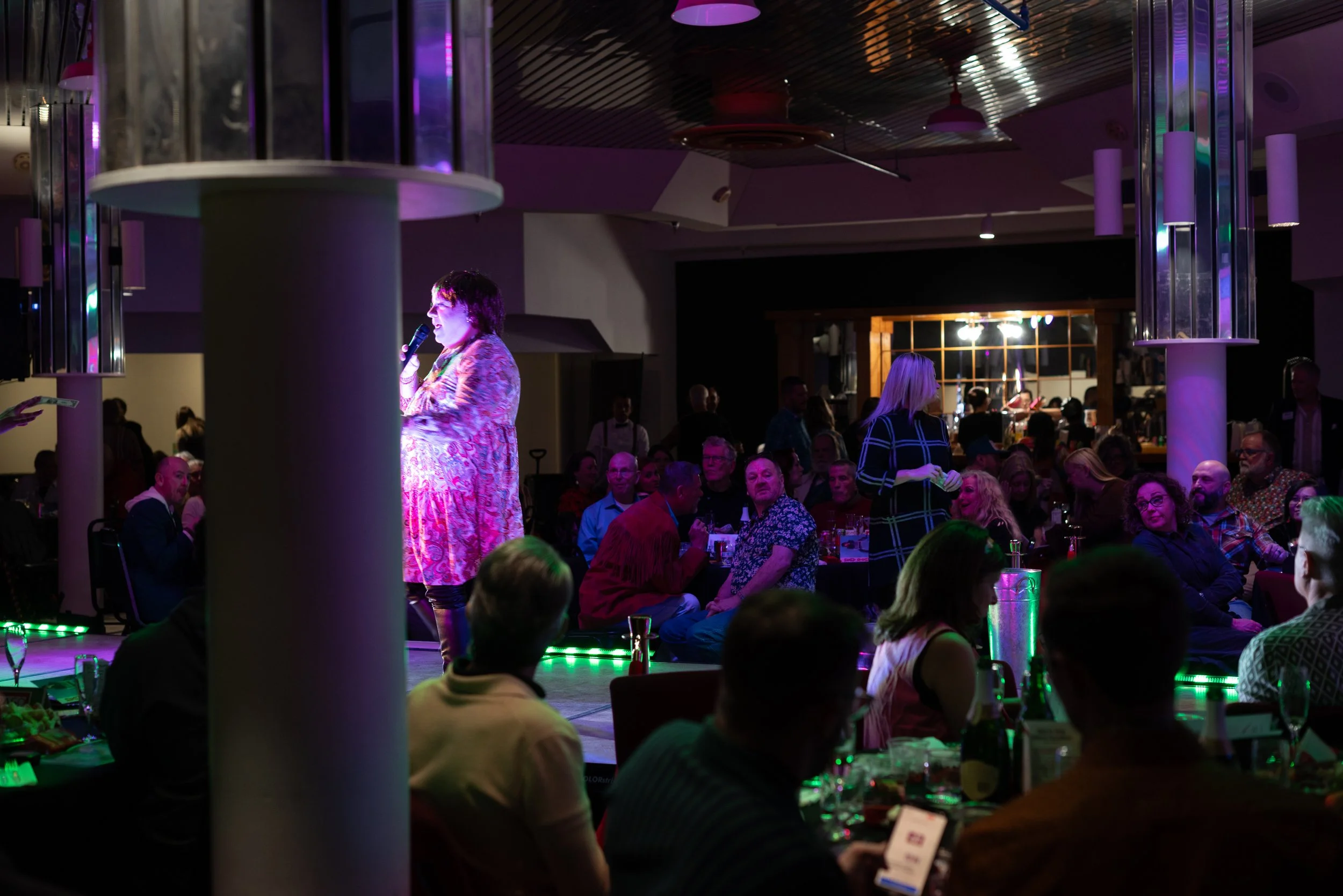 Woman on stage holding microphone, speaking or singing at a social event with seated audience in a dimly lit venue.