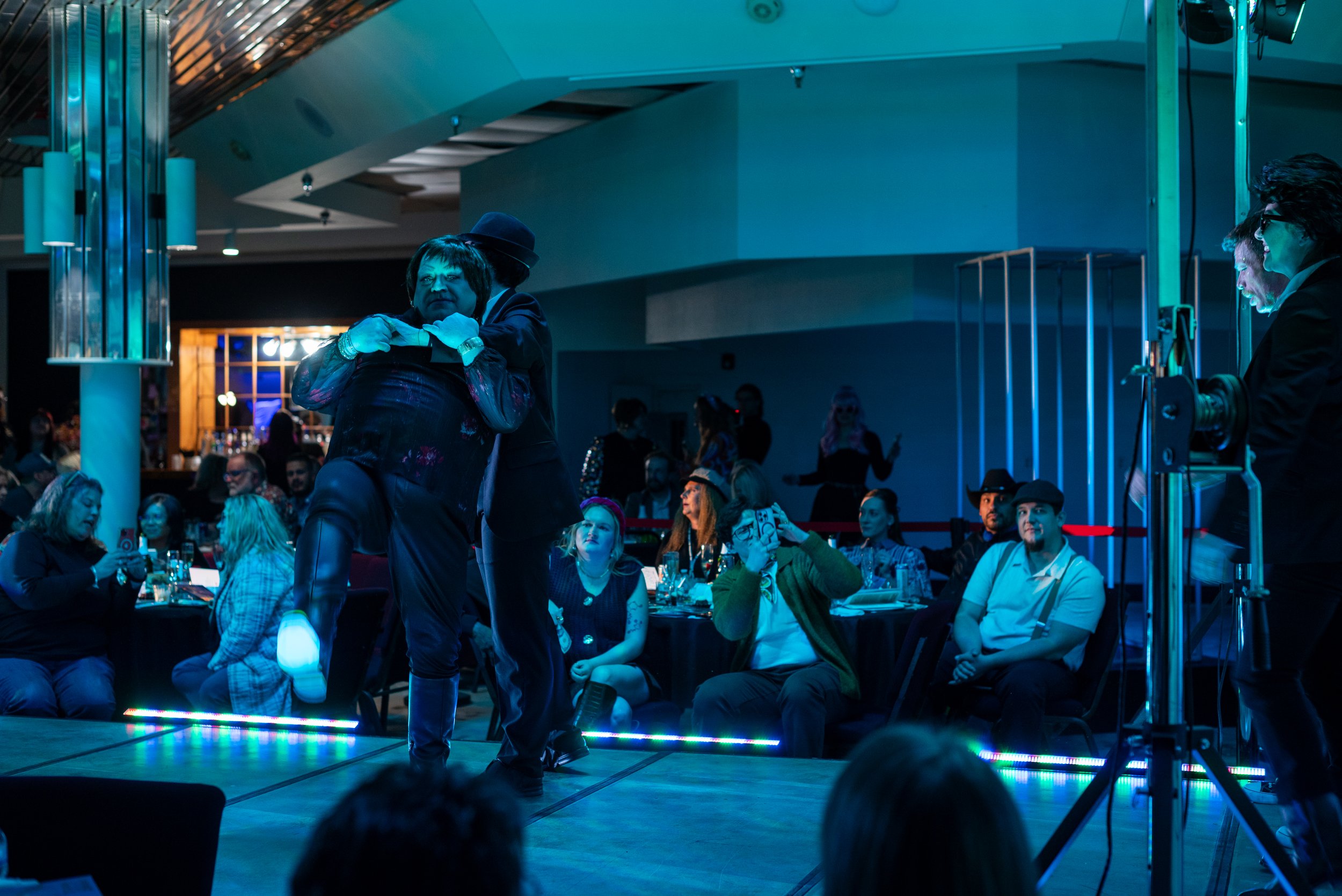 Couple dancing on stage at a lively event with an audience watching, some taking photos, in a dimly lit indoor setting with colorful lighting.