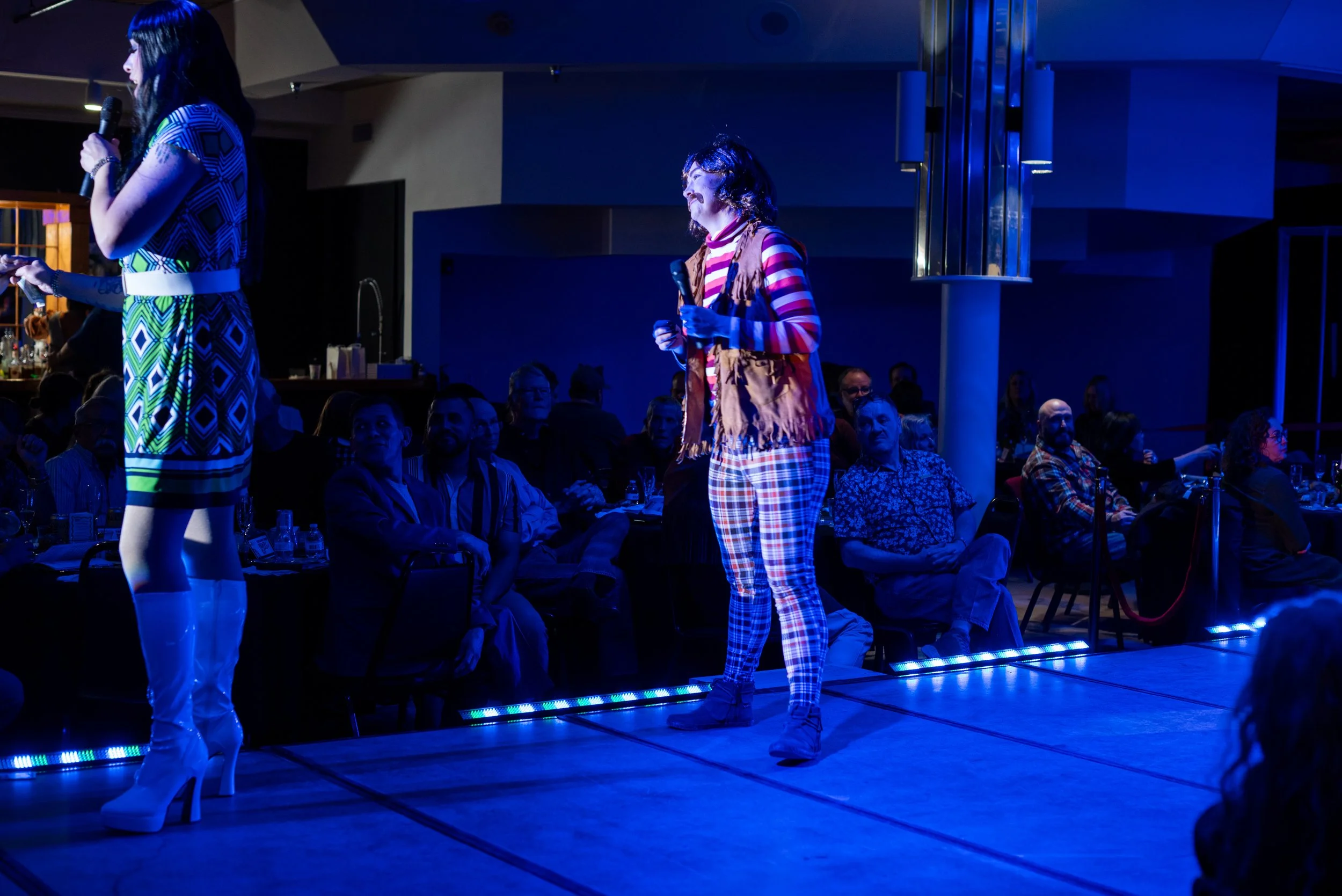 Two women stand on a stage under blue lighting, one speaking into a microphone and the other holding a microphone, with an audience seated in front watching them.