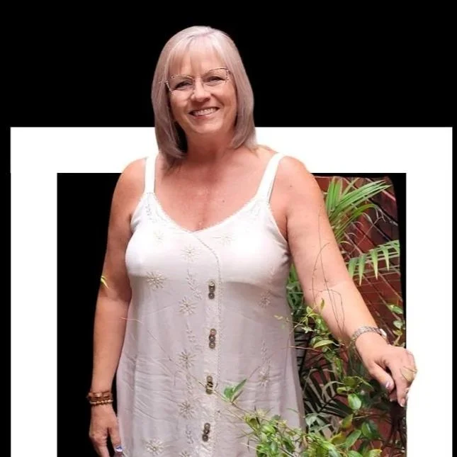 A smiling woman with shoulder-length gray hair and glasses, wearing a white sleeveless dress with embroidered flowers, standing outdoors beside a plant and a brick wall.