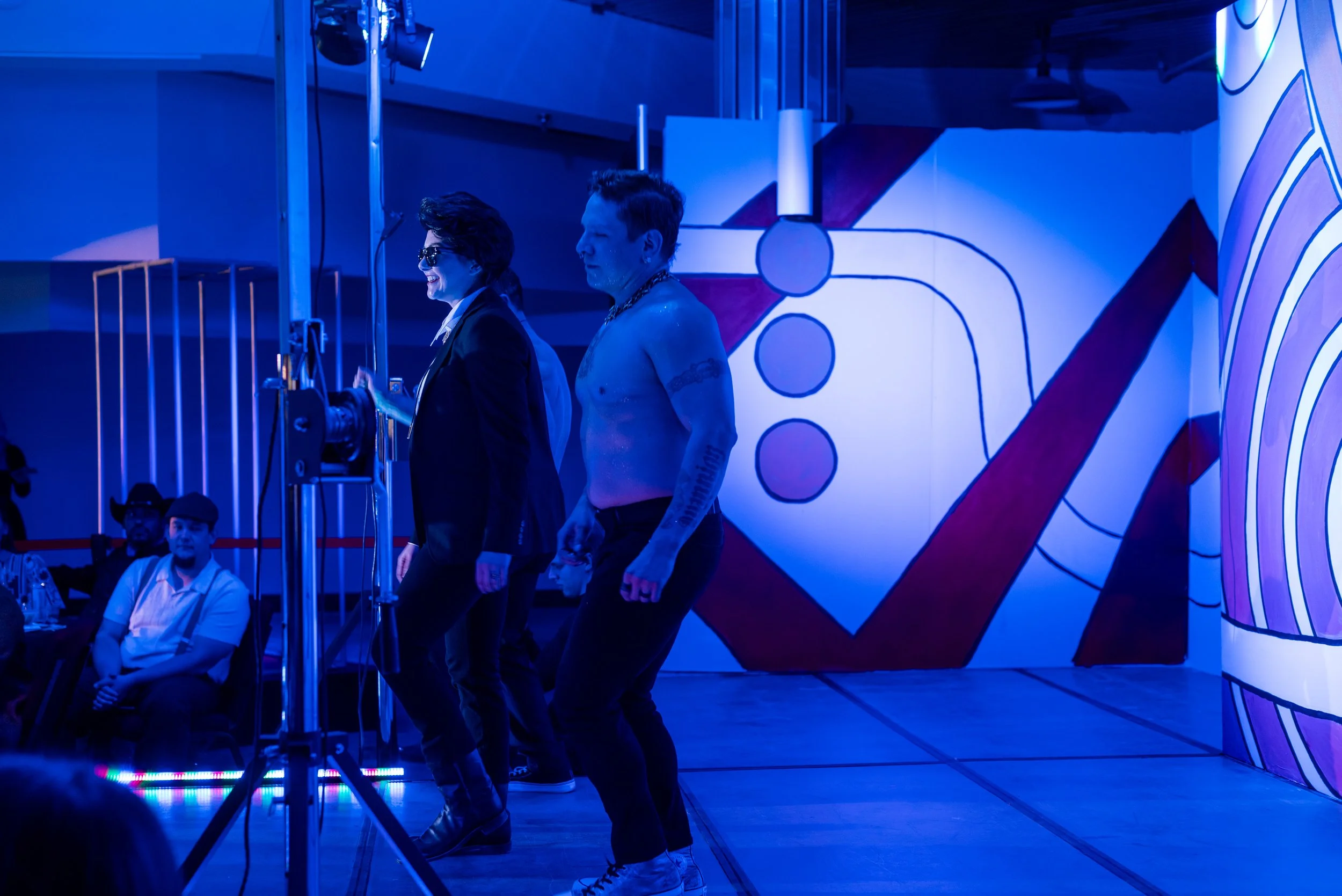 People dancing on stage in front of a colorful mural with abstract shapes, surrounded by blue lighting at an indoor event.