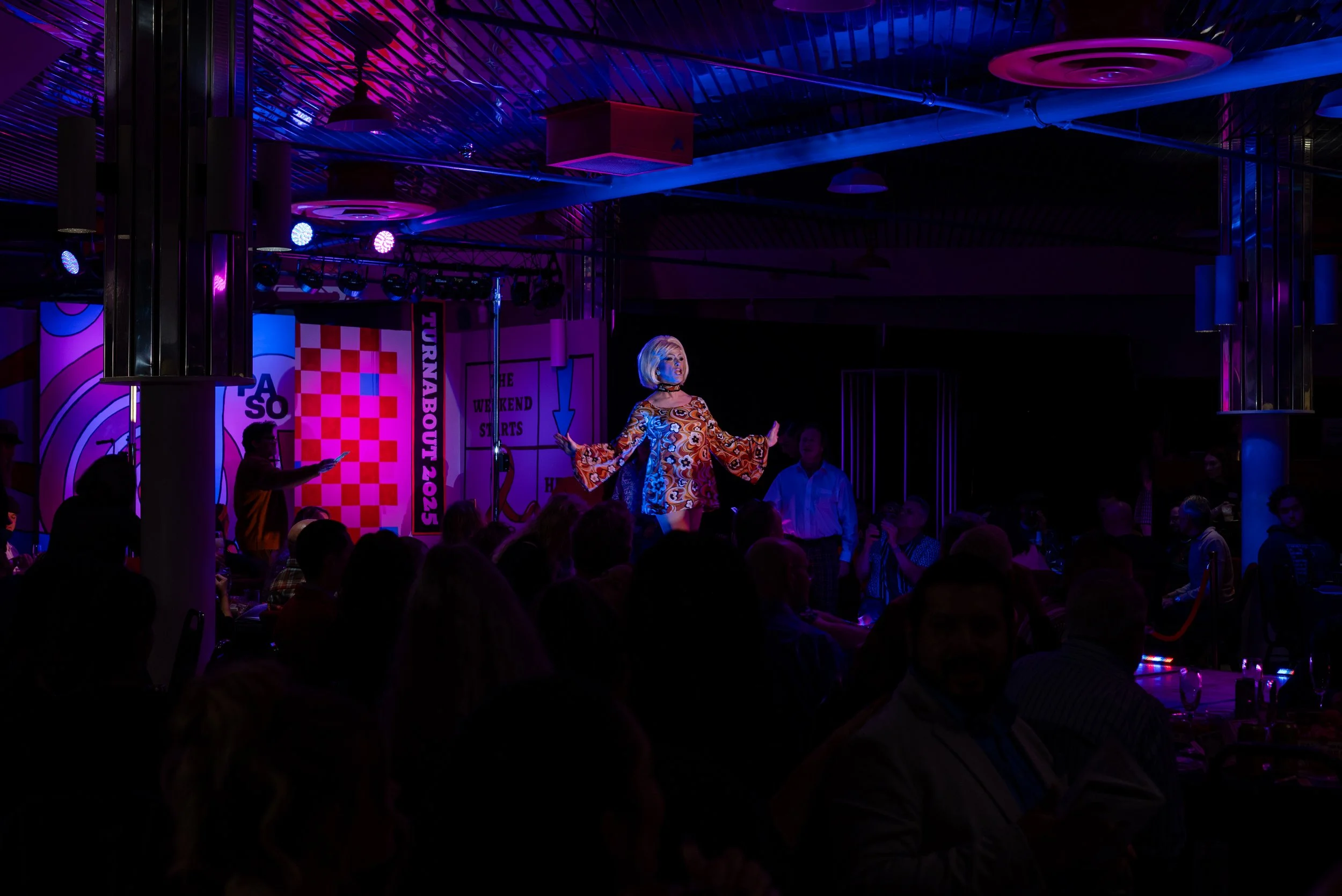 Performer on stage with colorful lighting and audience in a dimly lit venue.