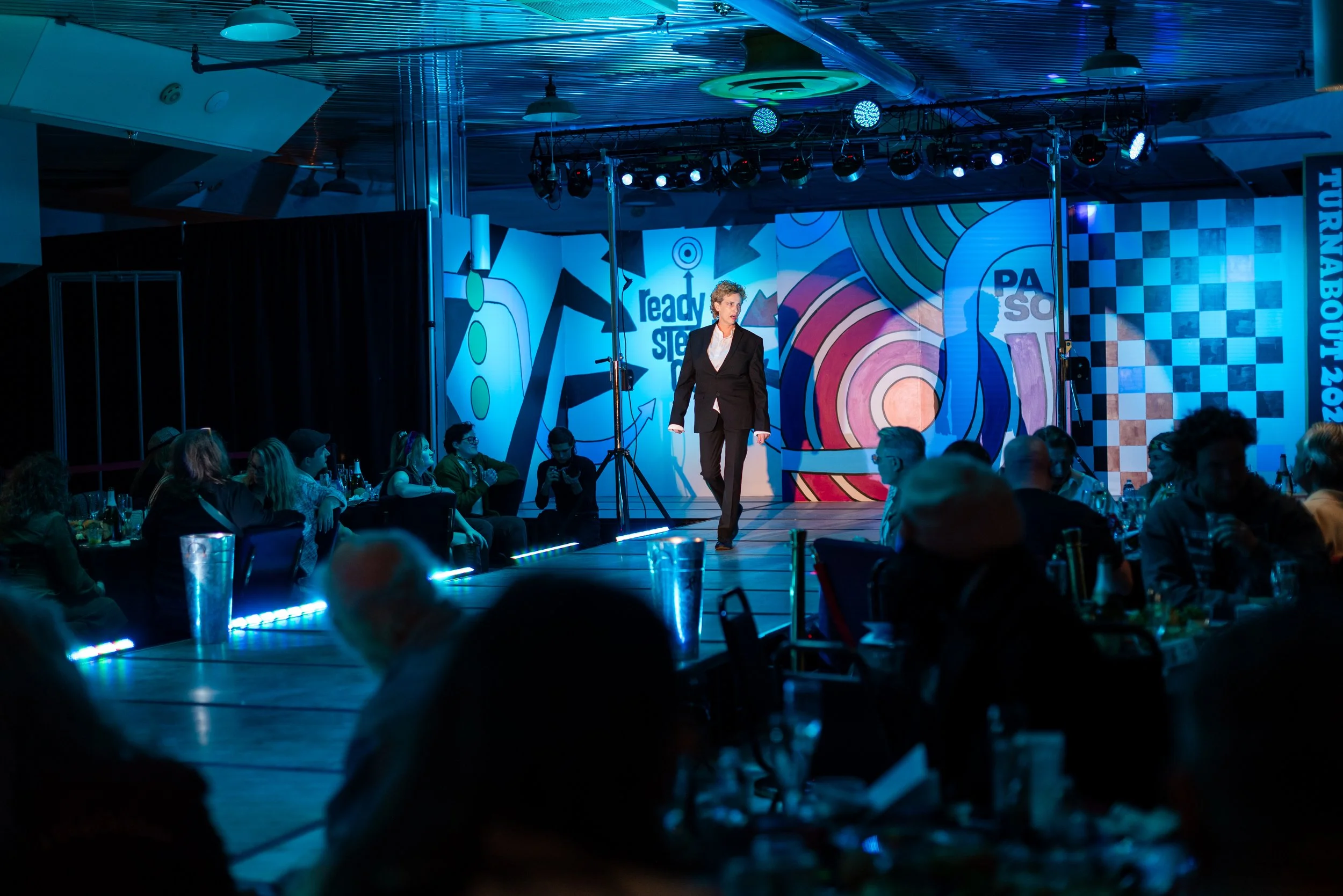 A person walking on stage during an event, with an audience seated at tables watching. The stage has colorful, abstract artwork in the background and stage lighting.