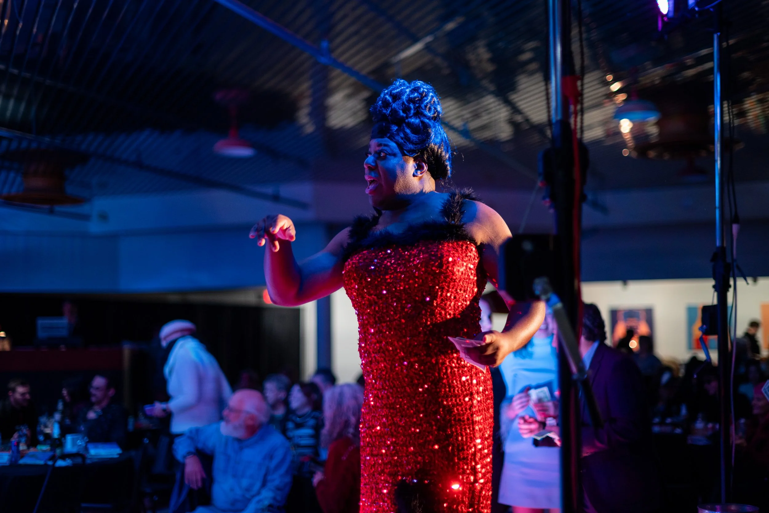 Performer in a red, sequined gown singing or speaking on stage at an event, with an audience seated at tables in the background.
