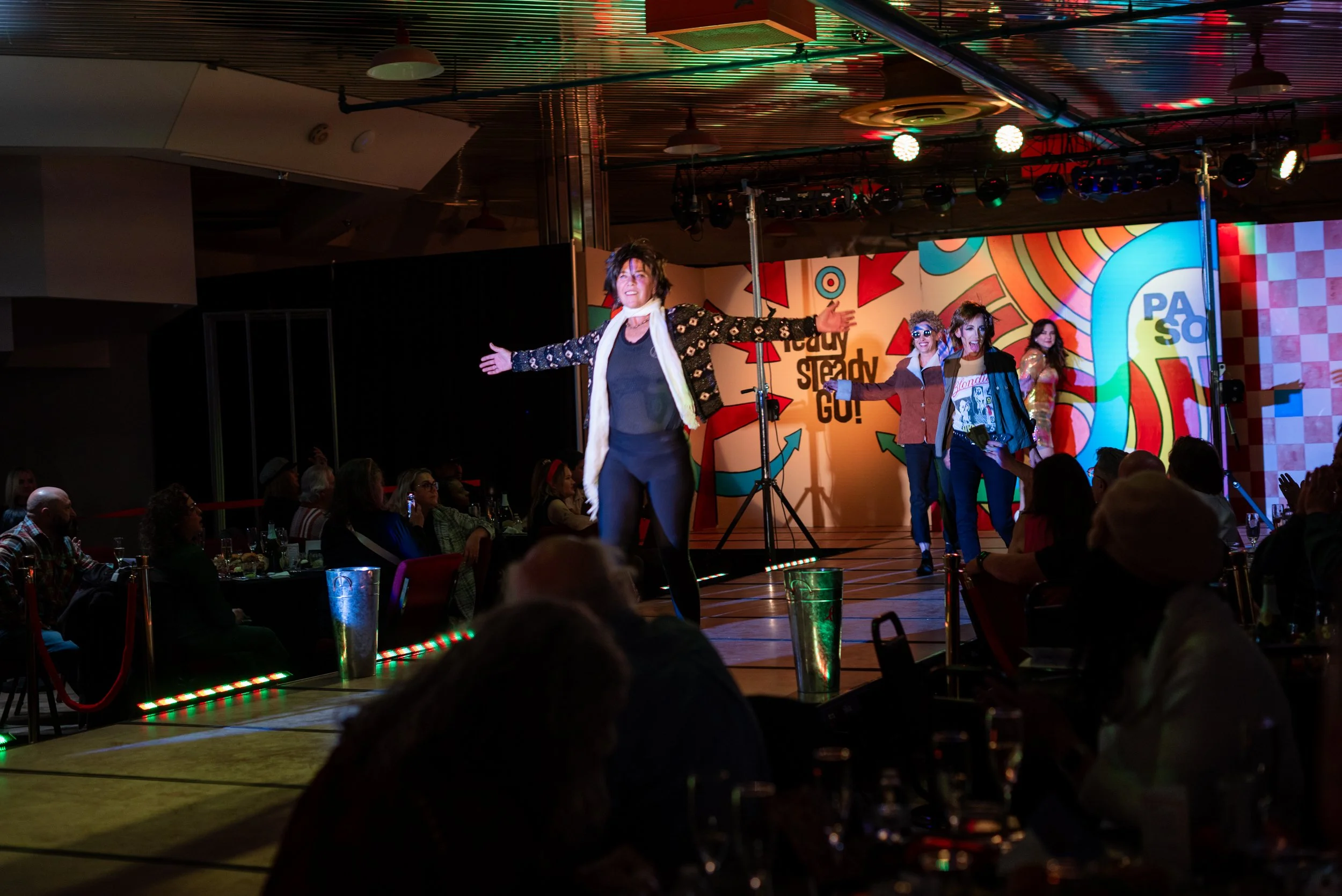 Performer on stage with arms wide open in front of colorful backdrop, with four other performers in the background and audience seated at tables watching.