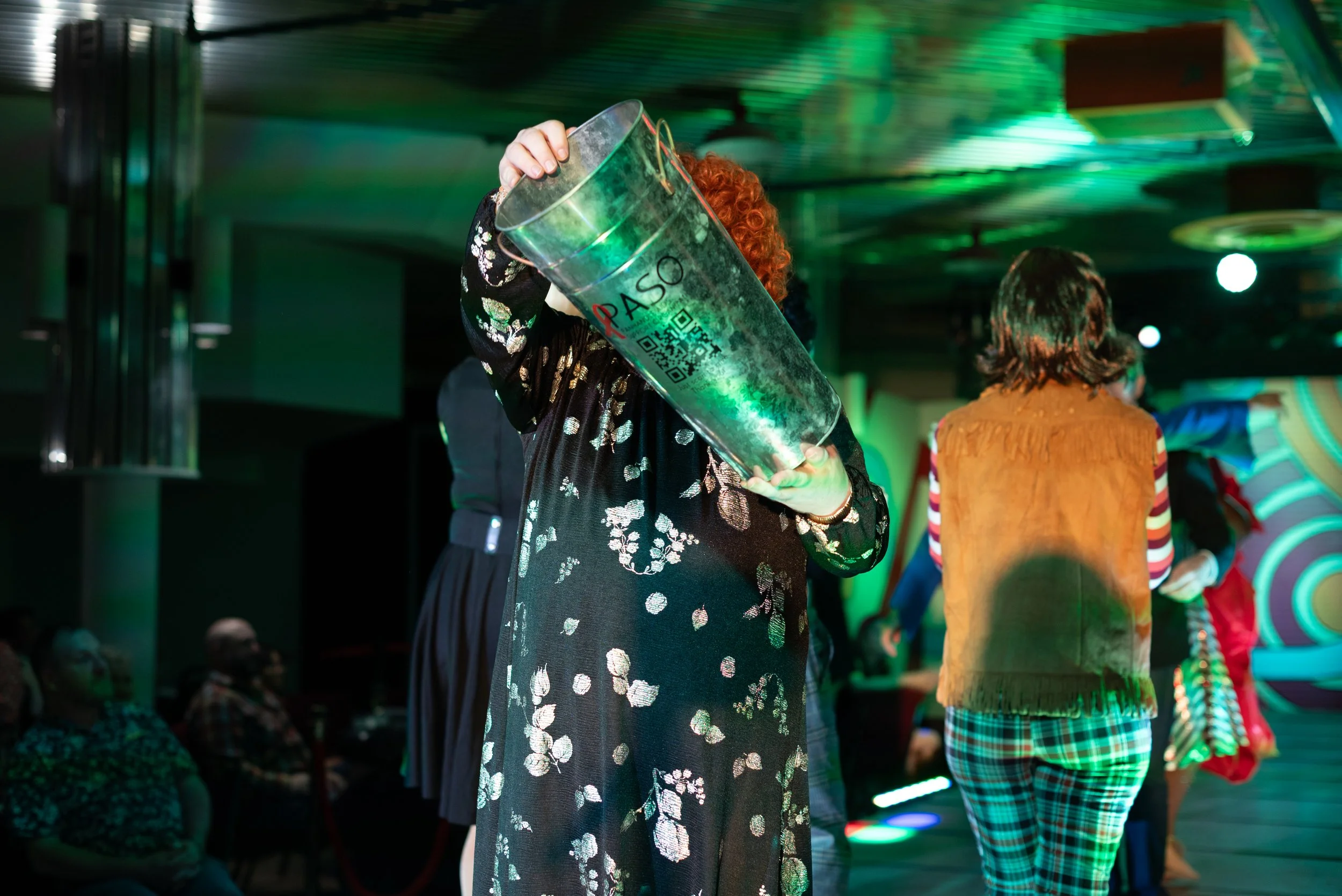 Person with curly red hair holding a large transparent container labeled 'PASO' with a QR code, at an indoor event with colorful lighting, other people in the background, and one person seated to the left.