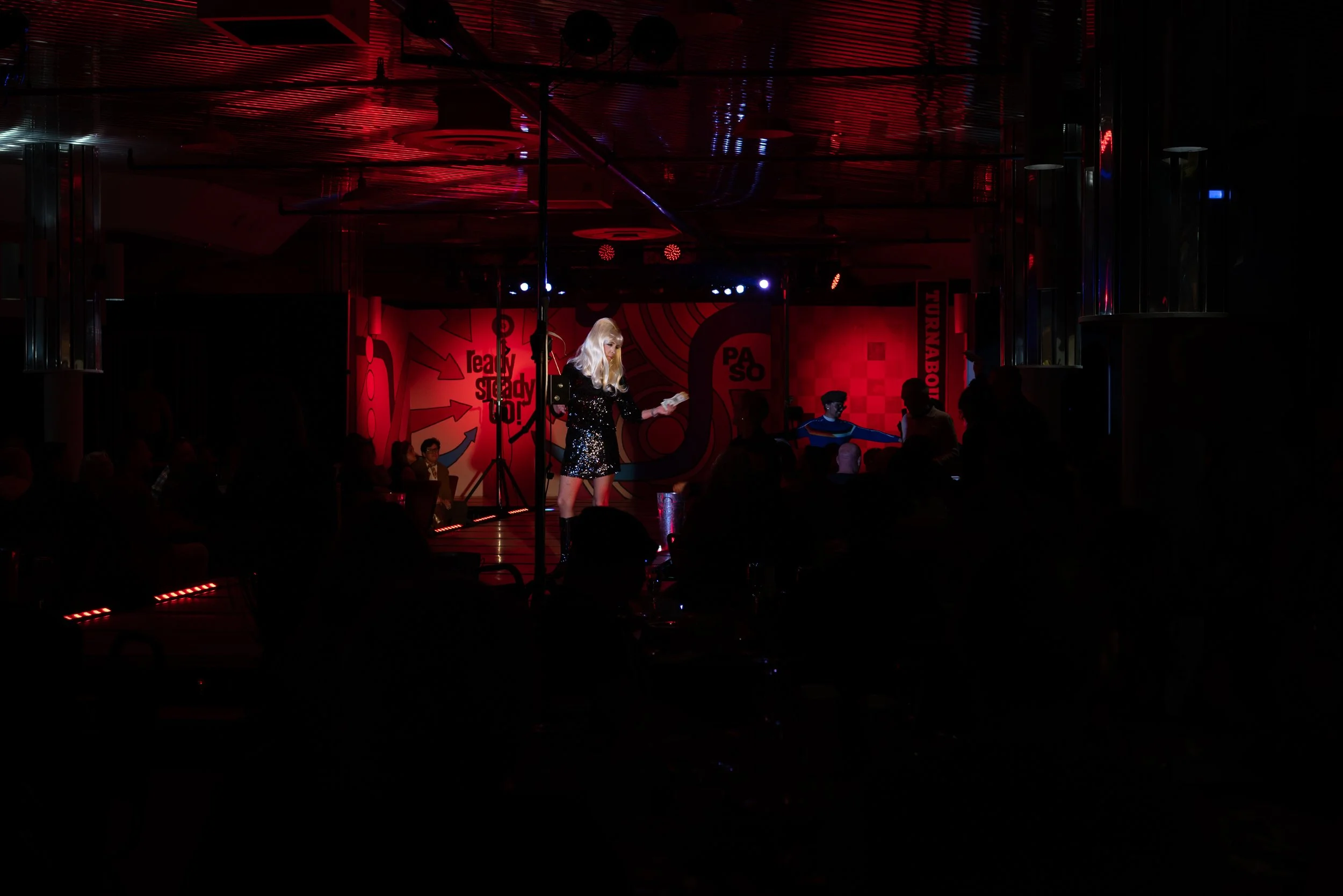 A woman with long blonde hair singing on a stage illuminated by red and blue lights, with a dark audience in front. The background features a colorful mural and various signs.