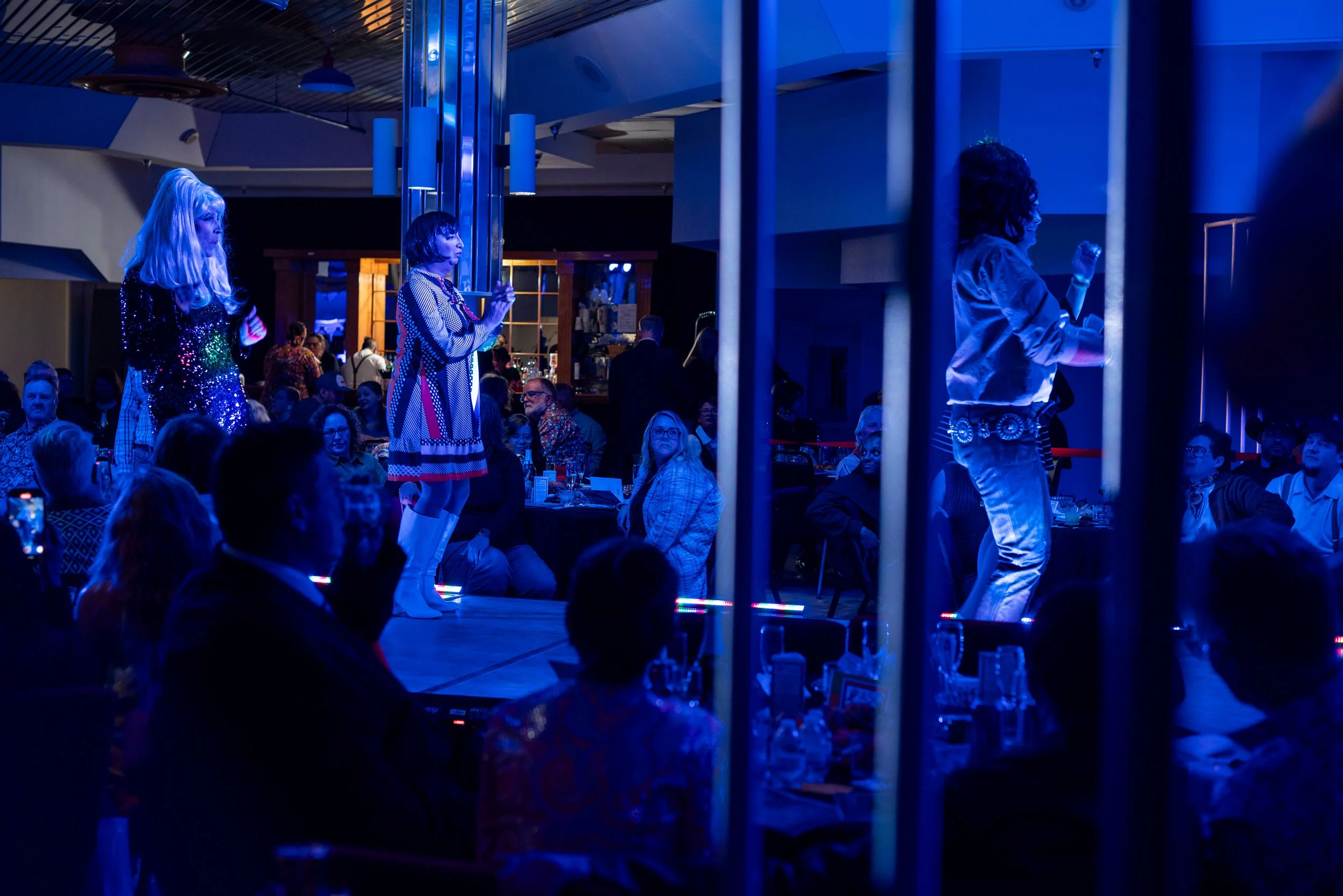 Three performers on a stage under blue lighting, two women and one man, dressed in shiny clothing, dancing or singing, with an audience seated at tables watching them.