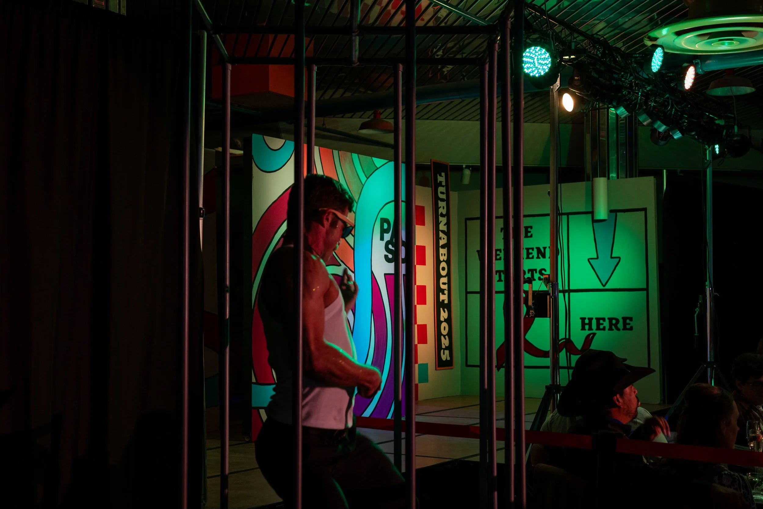 A person on stage wearing sunglasses, a white tank top, and dark pants, behind metal bars, with colorful signage in the background at a nightclub or bar event.