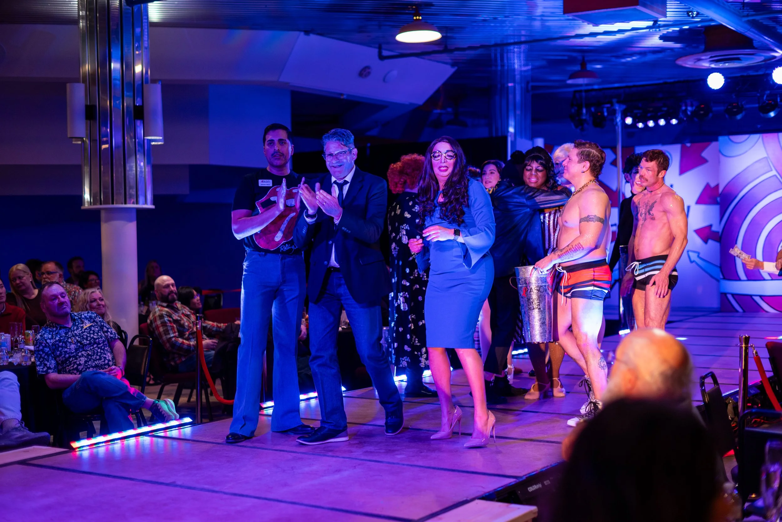 People on stage at a lively event with an audience watching, featuring diverse individuals in colorful and fashionable clothing.