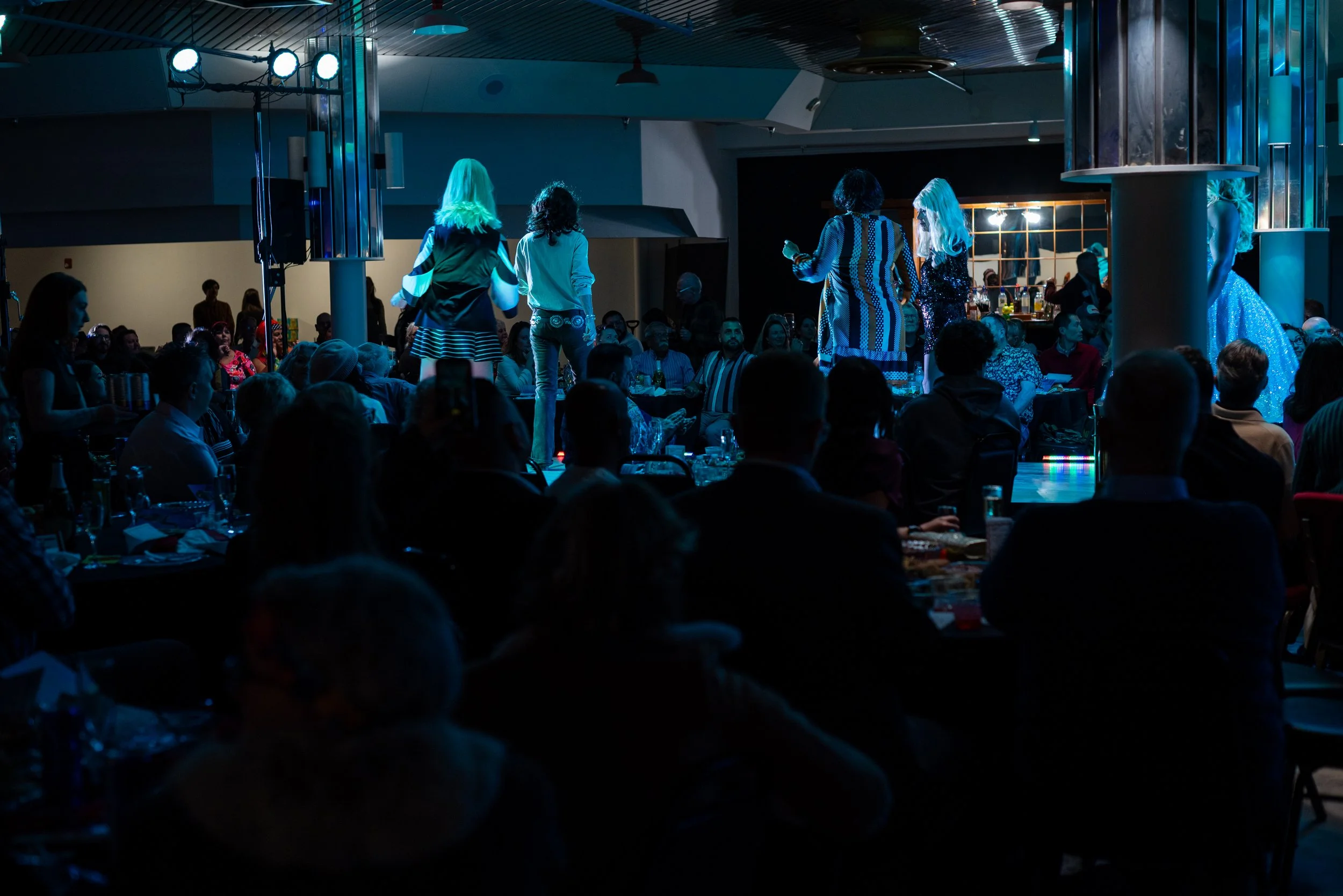 People watching a drag show on stage at a nightclub with colorful lighting and an audience seated at tables.