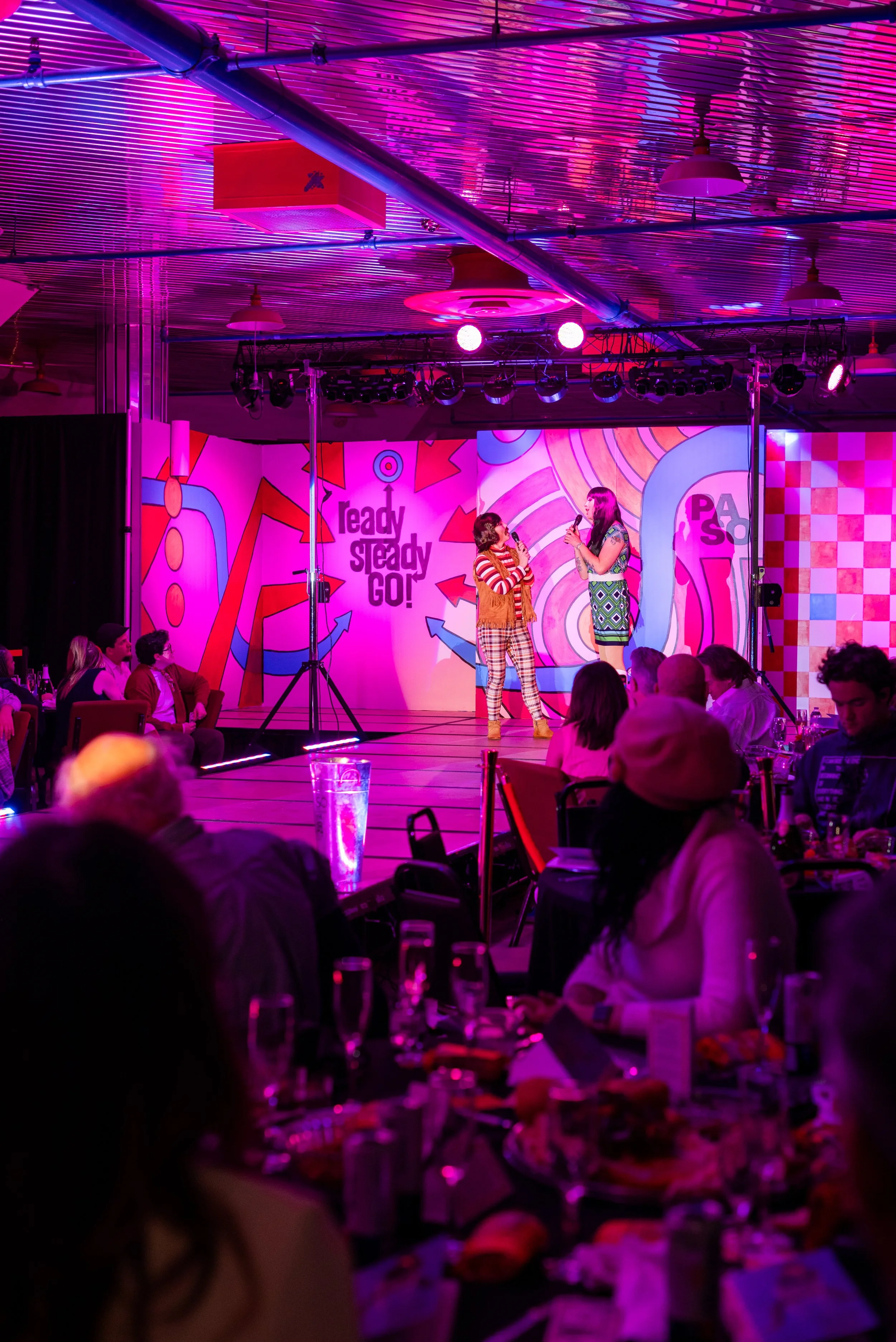 Two women stand on a brightly lit stage with colorful patterned backdrop, holding microphones, engaging in conversation. Audience seated at tables watch them, illuminated by vibrant pink and purple lighting.