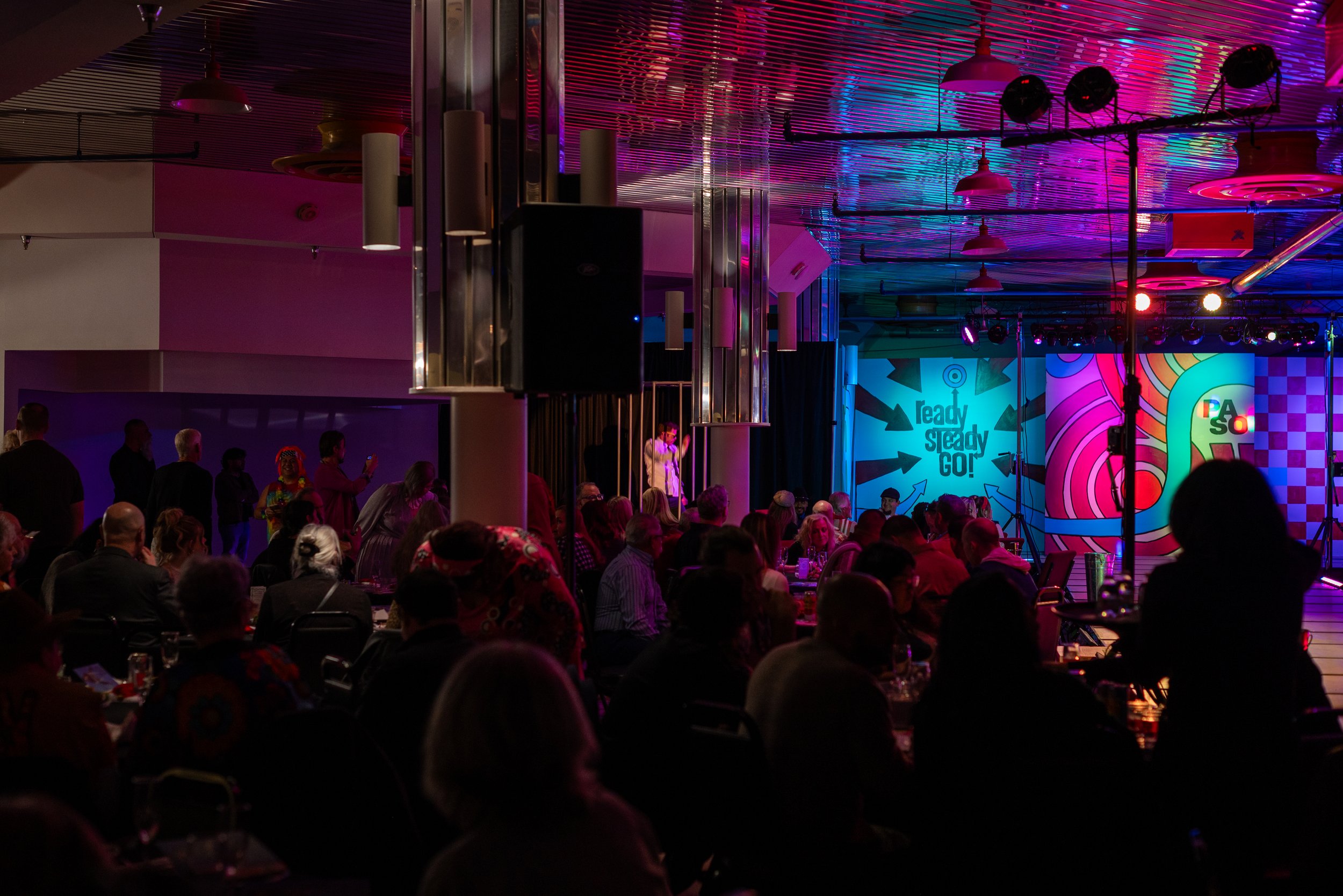 A crowded indoor event with people seated at tables, stage with colorful backdrop, and vibrant lighting.