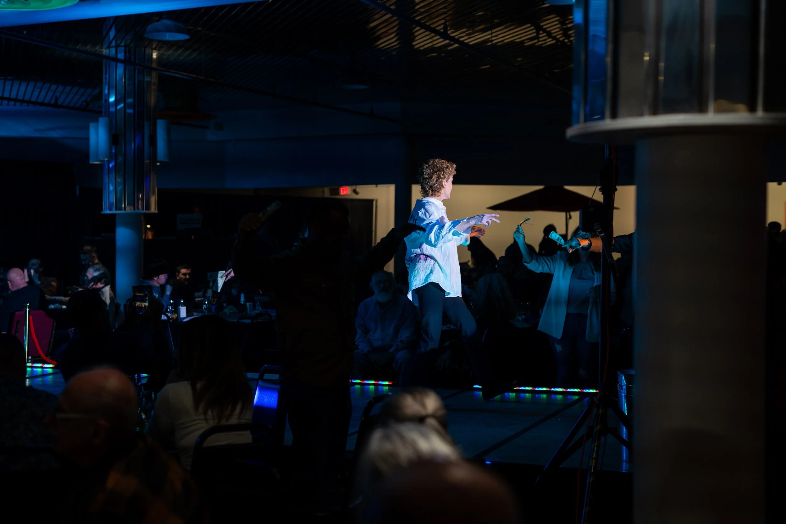 A person on stage appears to be dancing or performing, with a group of people sitting at tables in a dimly lit event space.