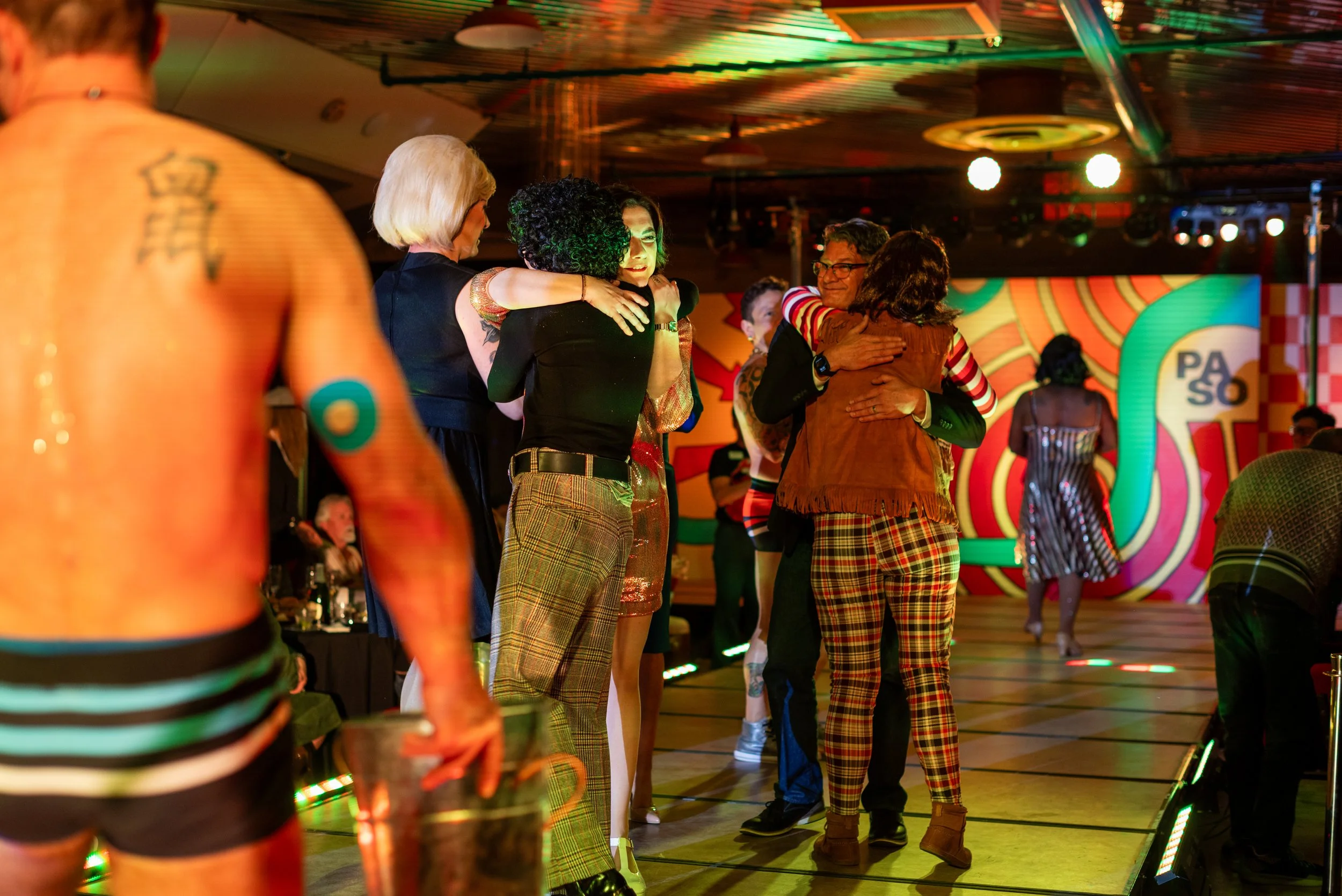 People dancing and hugging at a lively indoor nightclub or party with colorful lighting and a vibrant background that says PASO.
