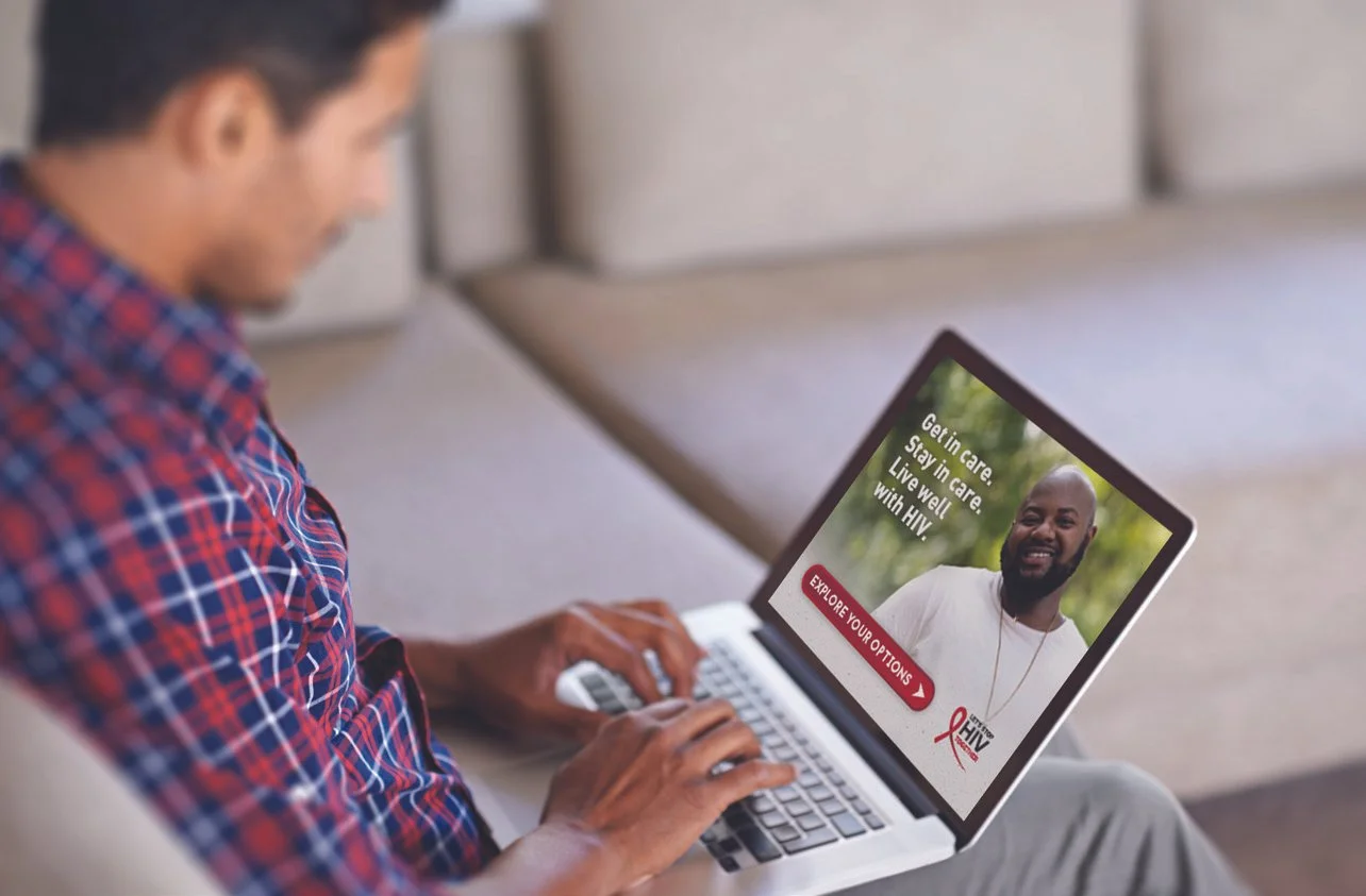 A man in a checkered shirt sitting on a couch using a laptop to view an HIV awareness webpage, which features a smiling man and messages about HIV care and prevention.