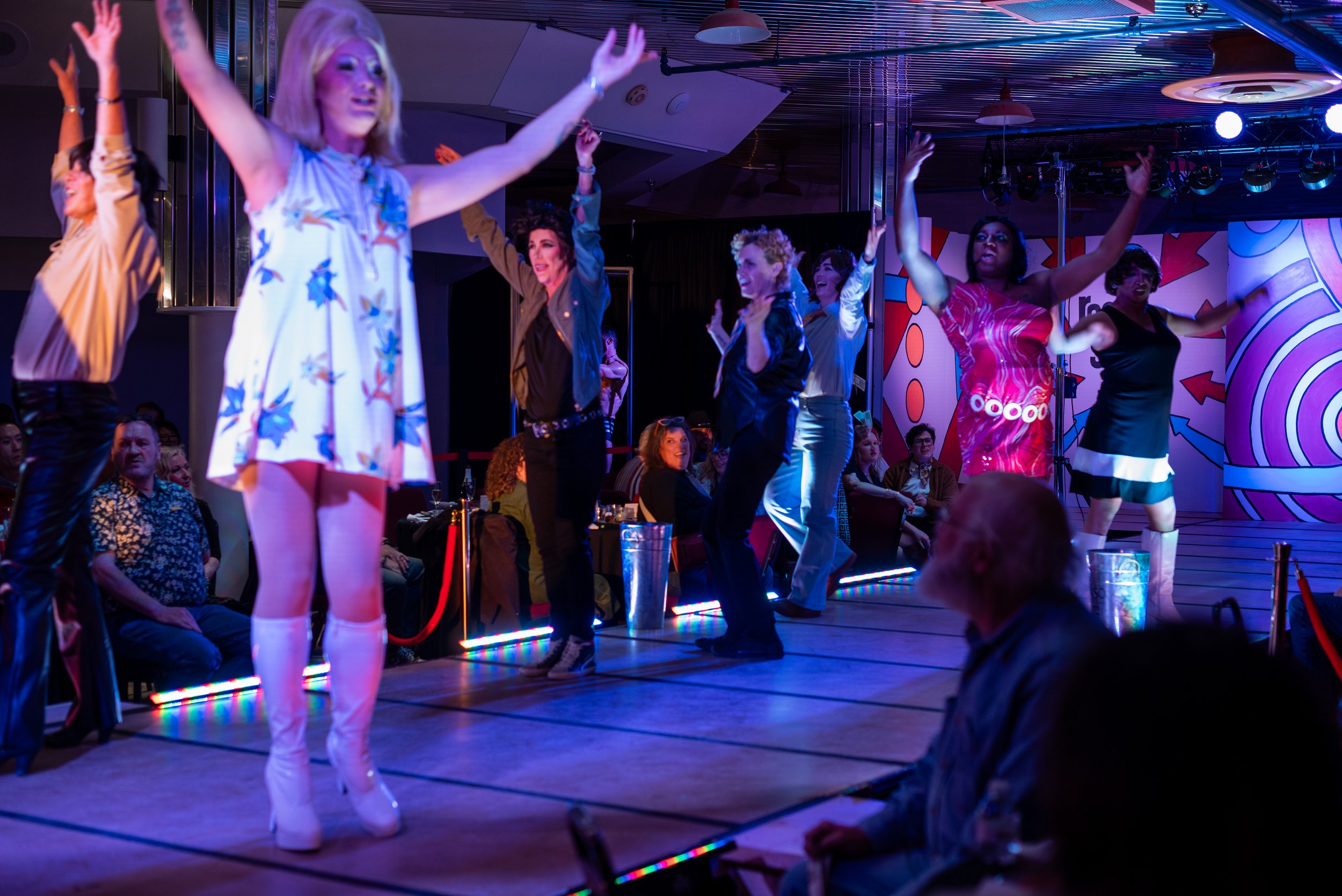 People dancing on a stage with colorful background lights and an audience watching.
