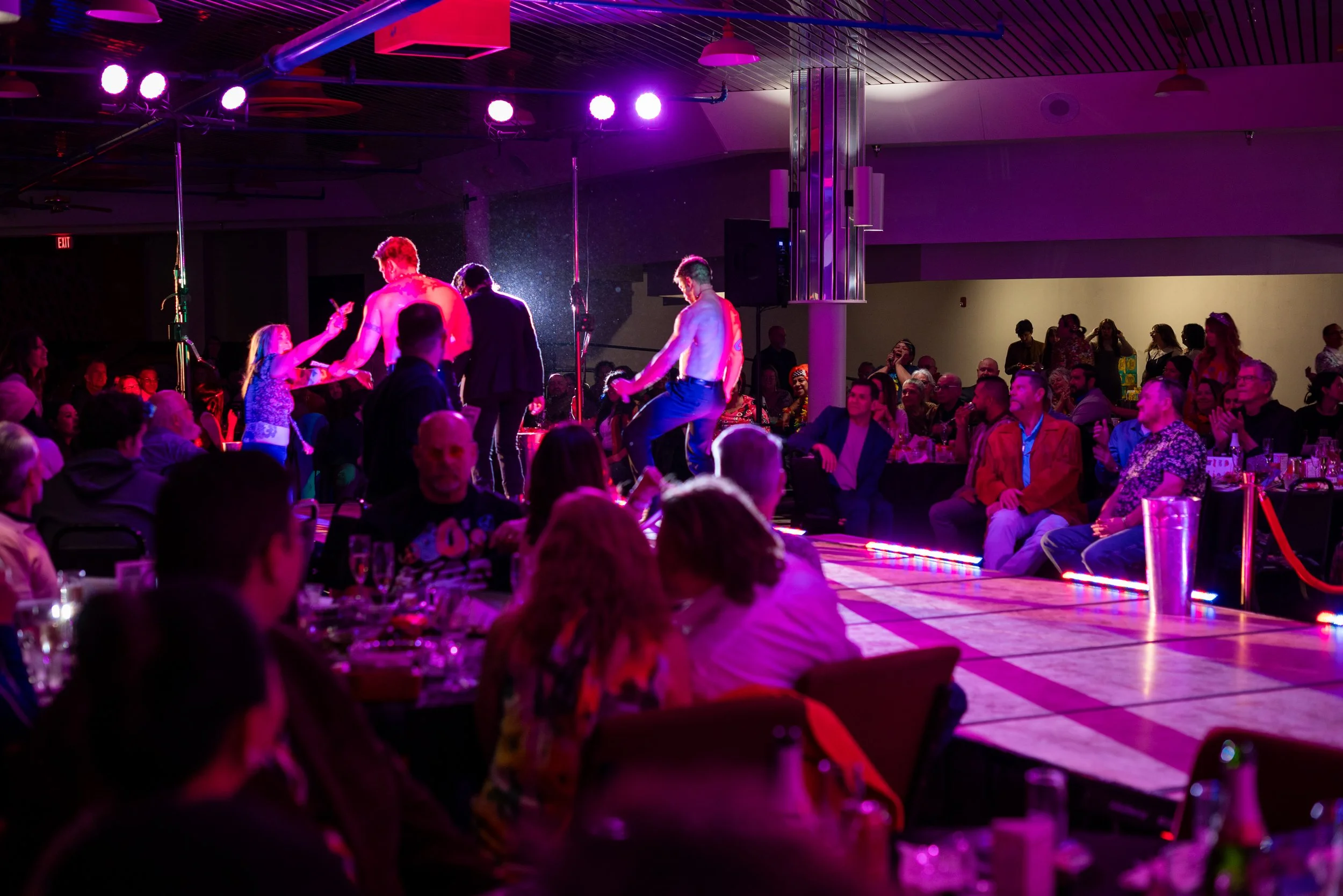 People watching a drag show on a stage with purple and pink neon lights in a nightclub.