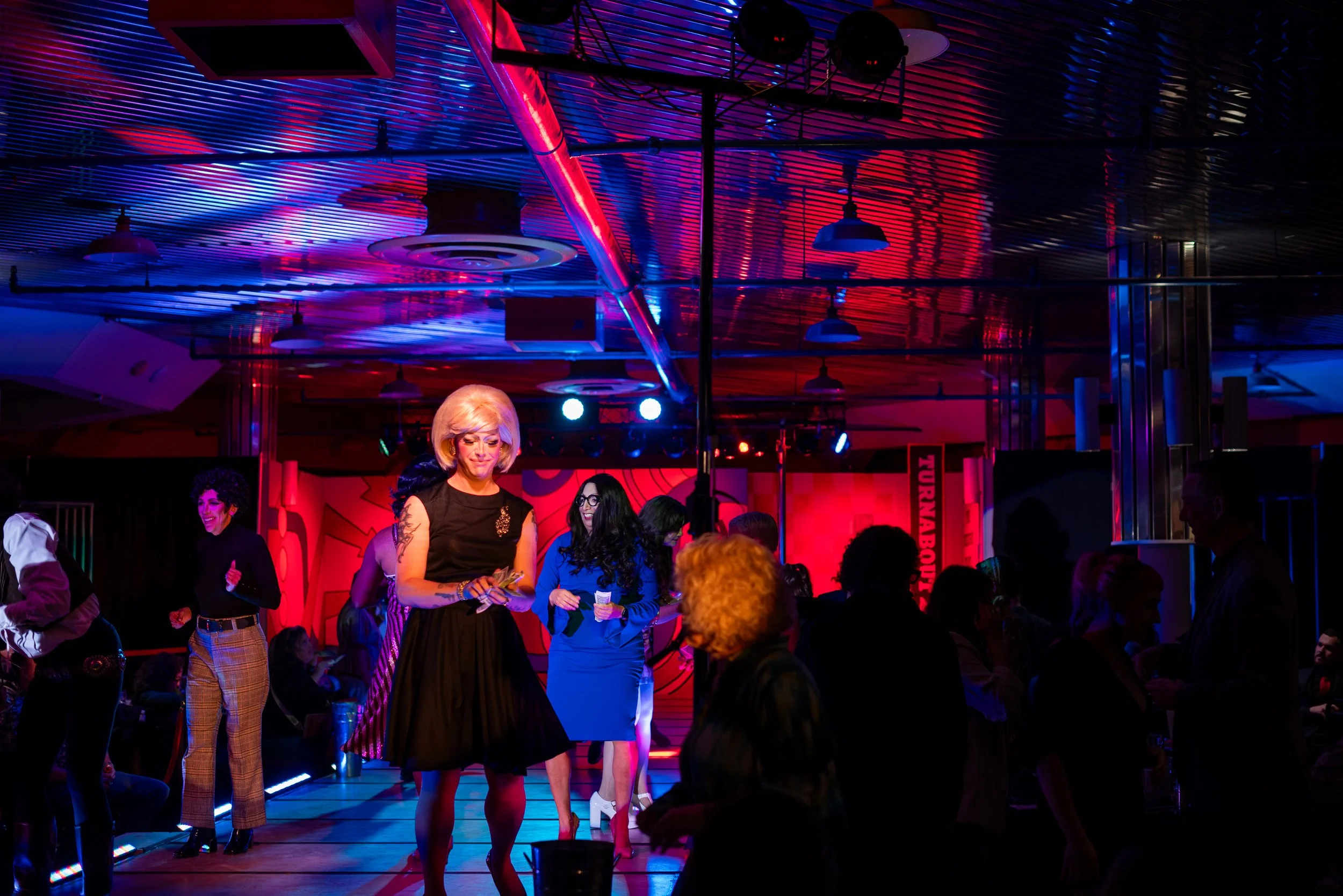 People dancing and socializing at a vibrant nightclub with colorful neon lights and a reflective ceiling.