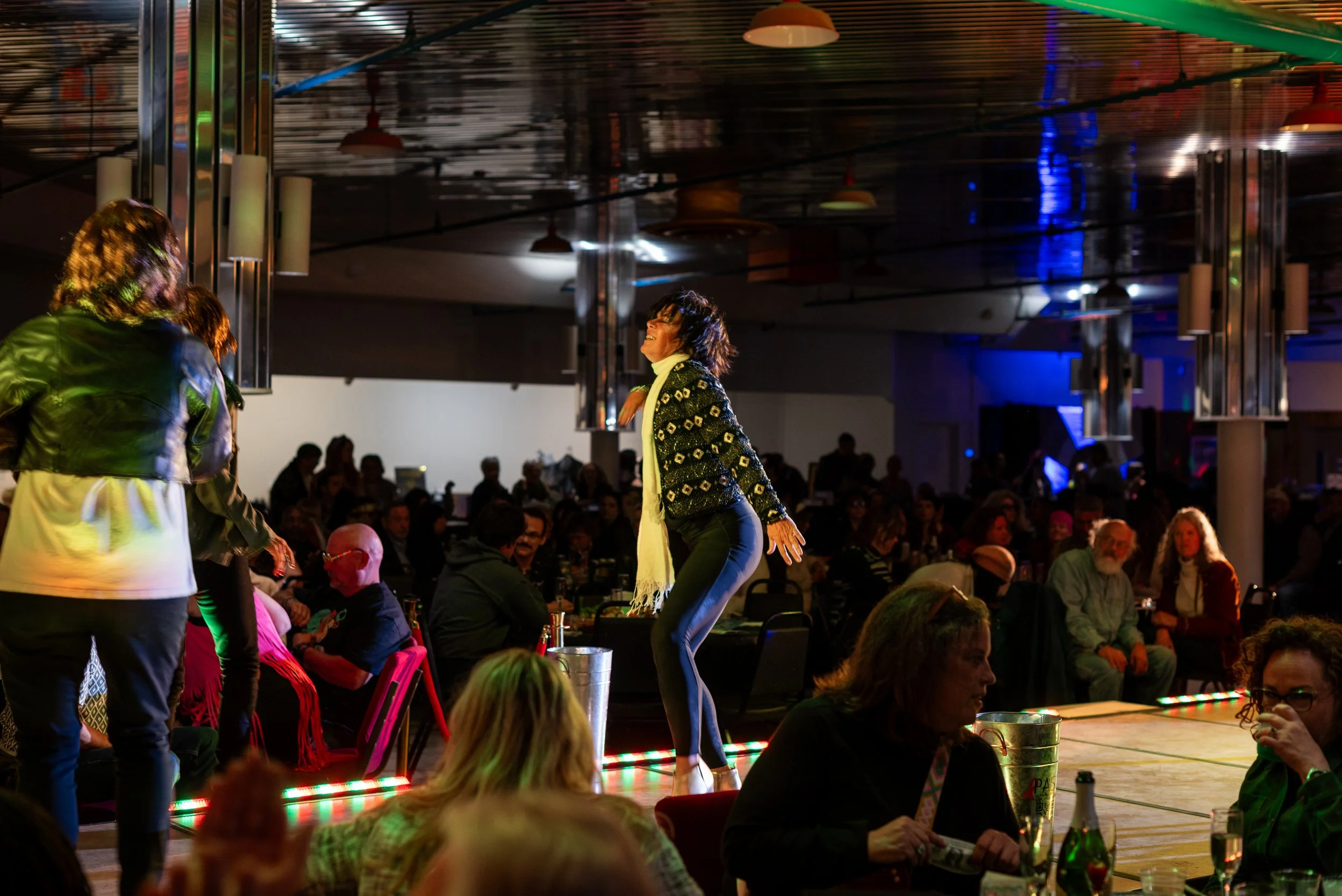 A woman dancing on a stage at a lively indoor event with a crowd seated around. The woman is wearing a patterned blazer, dark pants, and a scarf, smiling as she dances. The atmosphere is vibrant with colorful lighting and decorations.