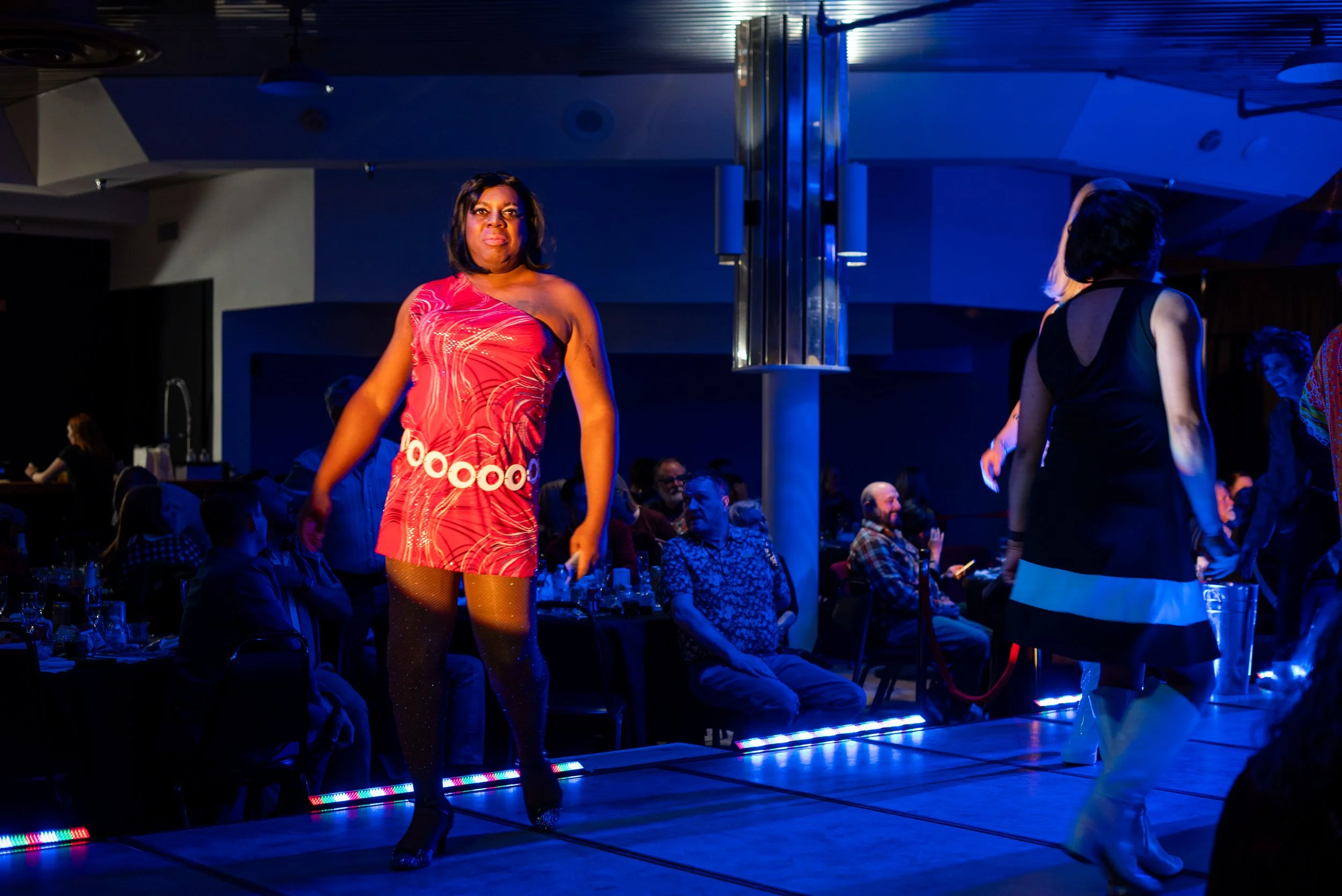 Two women dancing on a stage with an audience watching, colorful lighting, and a modern interior setting.