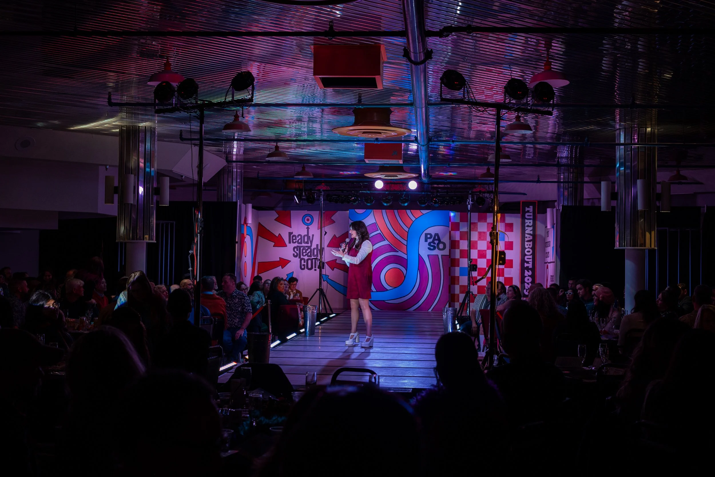 A comedian performing on stage at a comedy show event, with an audience seated at tables in a dimly lit venue with colorful backdrop.