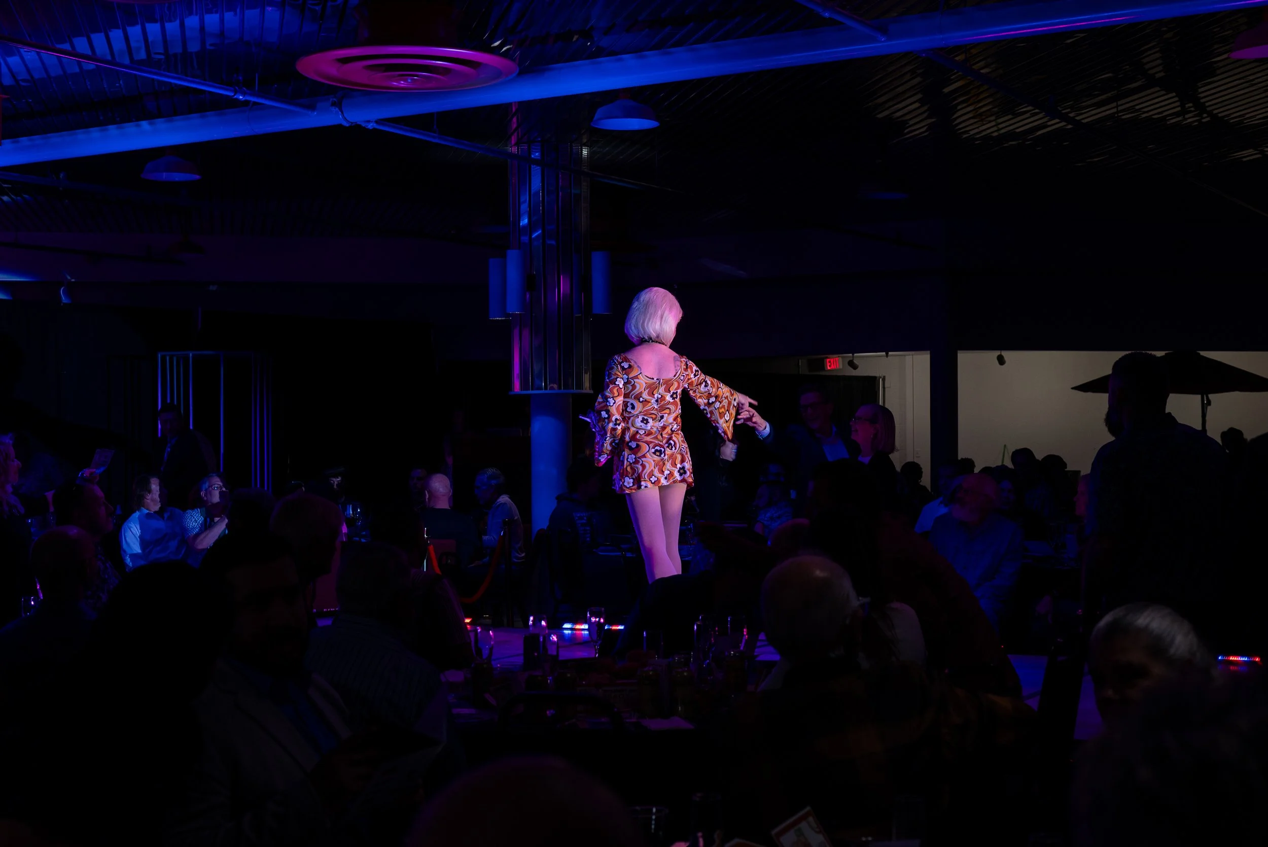 A woman with short blonde hair stands on a stage in a dimly lit venue, wearing a colorful patterned dress, surrounded by an audience seated at tables.