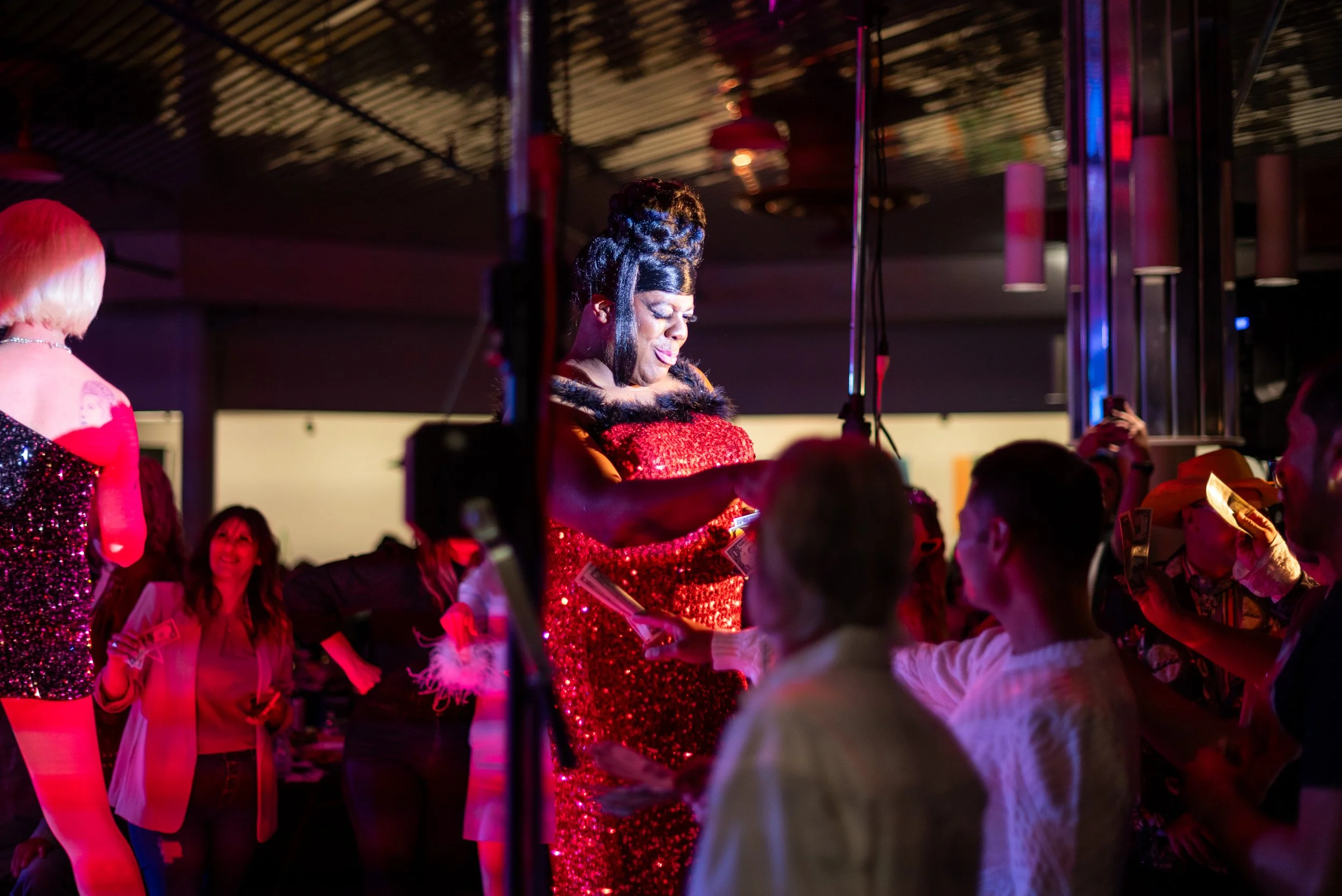 A woman in a red sequin dress is on stage at a nightclub, receiving a large sum of cash from audience members. The scene is vibrant with colorful lighting and other people visible in the background.