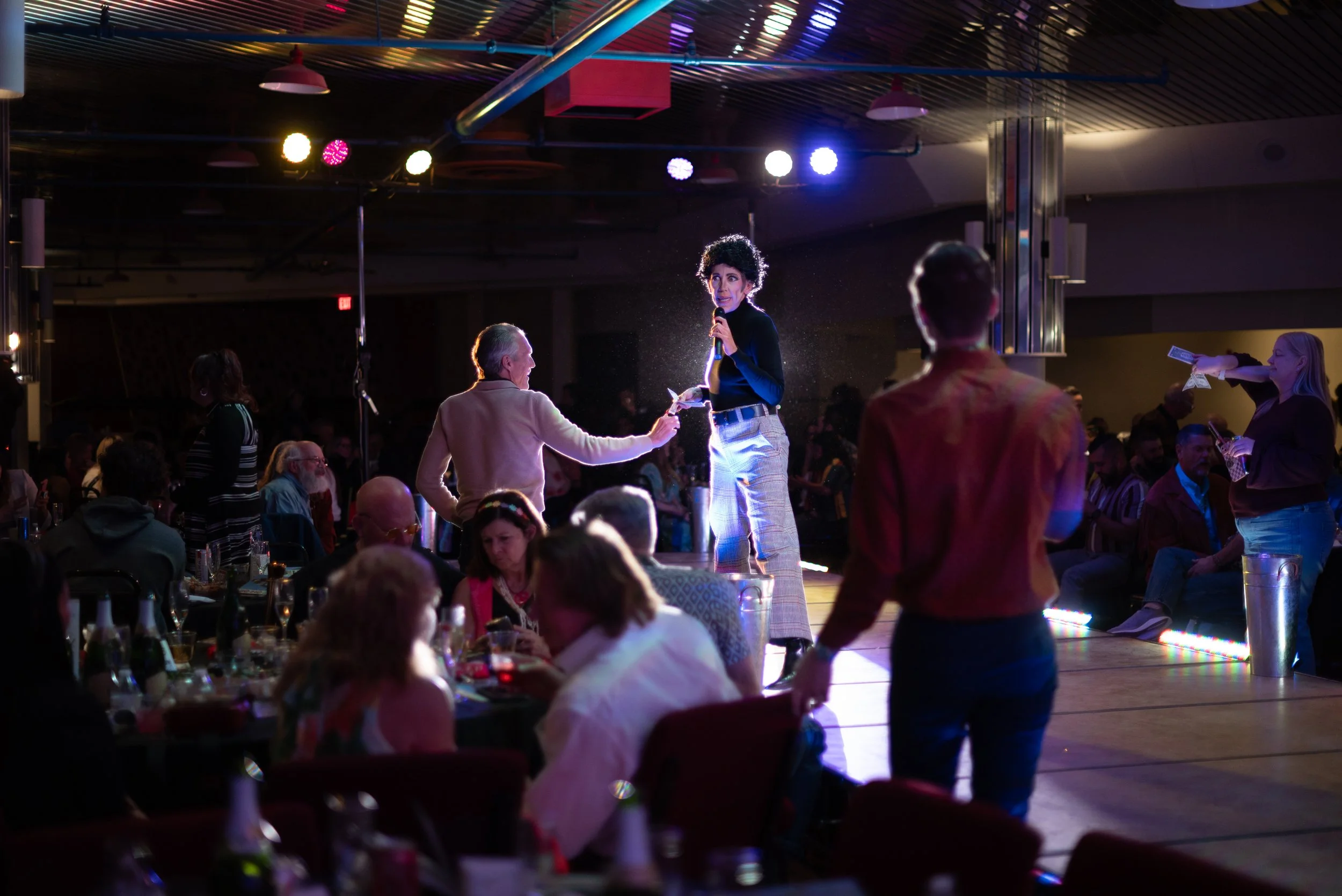 A woman on stage holding a microphone, with one hand extended towards a person who is handing her money, surrounded by an audience seated at tables, at an event or performance in a dimly lit venue.