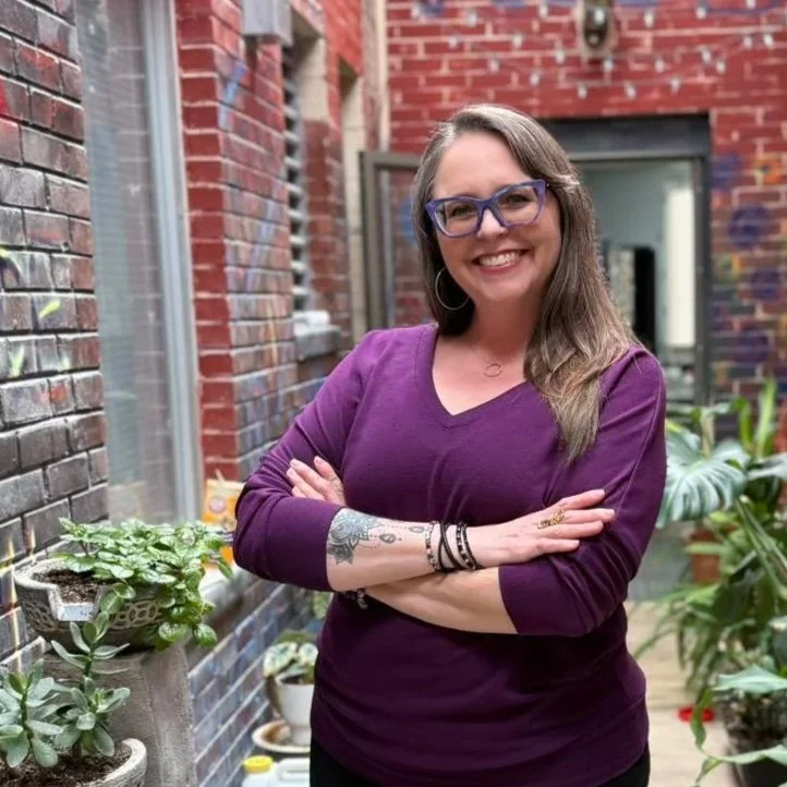 Smiling woman with long brown hair, wearing glasses and a purple top, standing with arms crossed in an outdoor brick patio with plants.