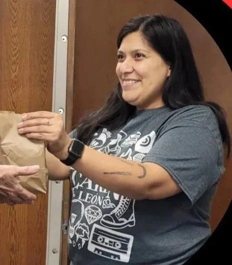 A woman with dark hair smiling as she hands a brown paper bag to someone. She is wearing a gray T-shirt with white graphics and a black smartwatch on her left wrist.