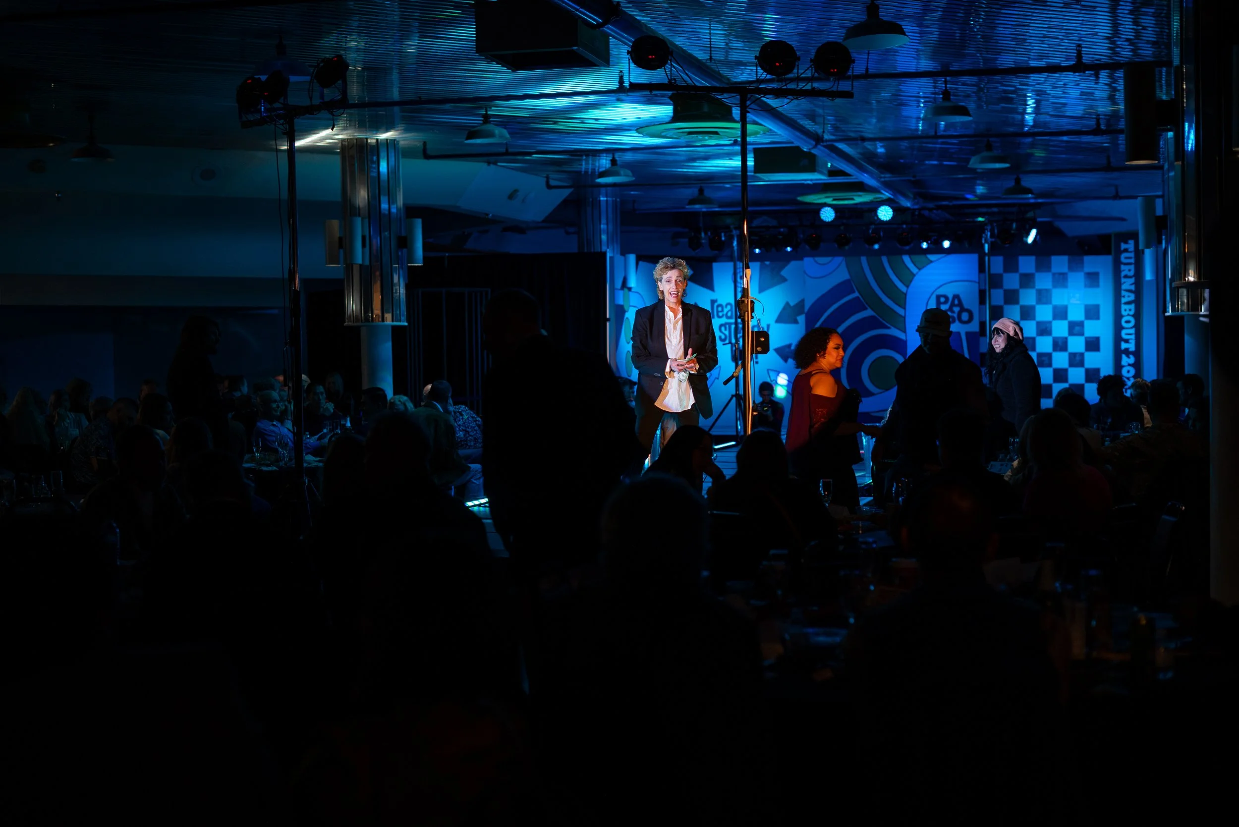 A woman in business attire stands on stage with a microphone, illuminated by blue and white lights, while an audience watches in a dimly lit conference or event room with a backdrop displaying the words 'Turnabout 2023' and a checkered pattern.