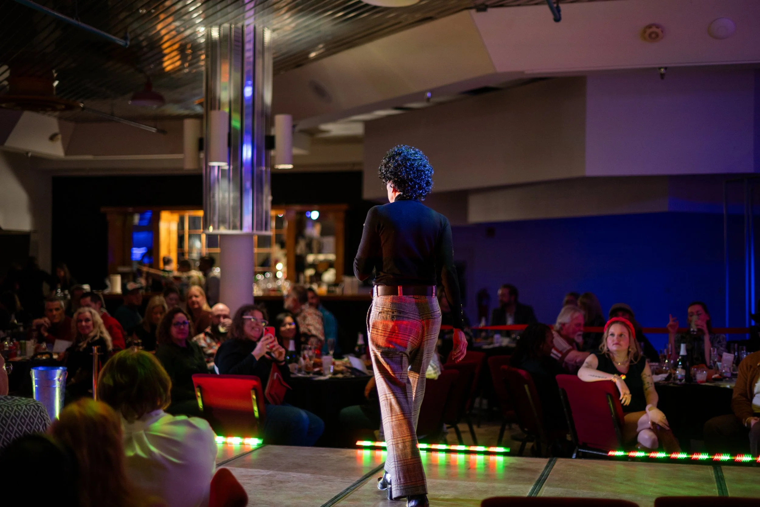 Person performing on stage in front of an audience in a dimly lit venue with colorful lighting.