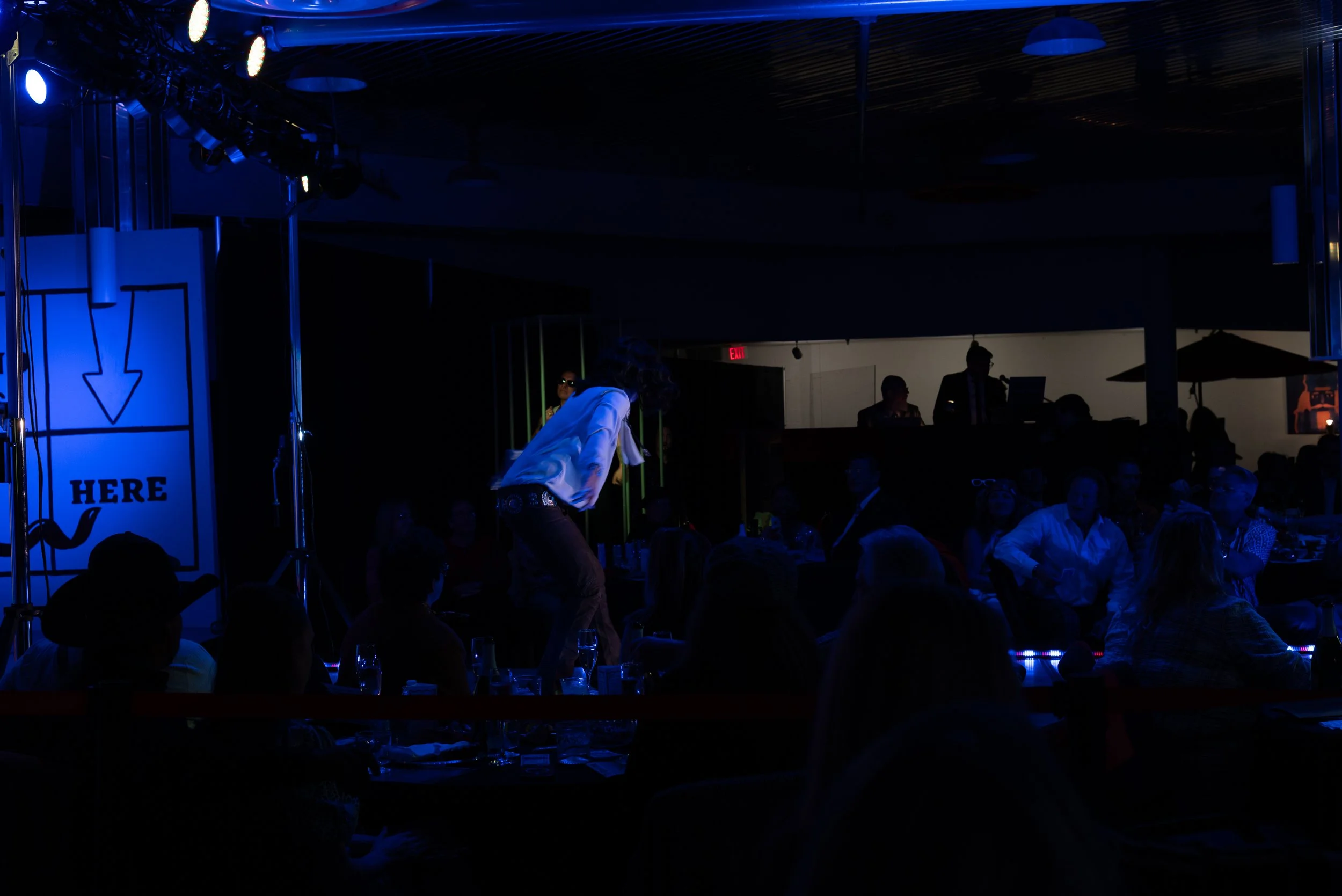 Performer on stage with audience in a dimly lit venue, blue lighting, sign with arrow and 'HERE' text visible on the left.
