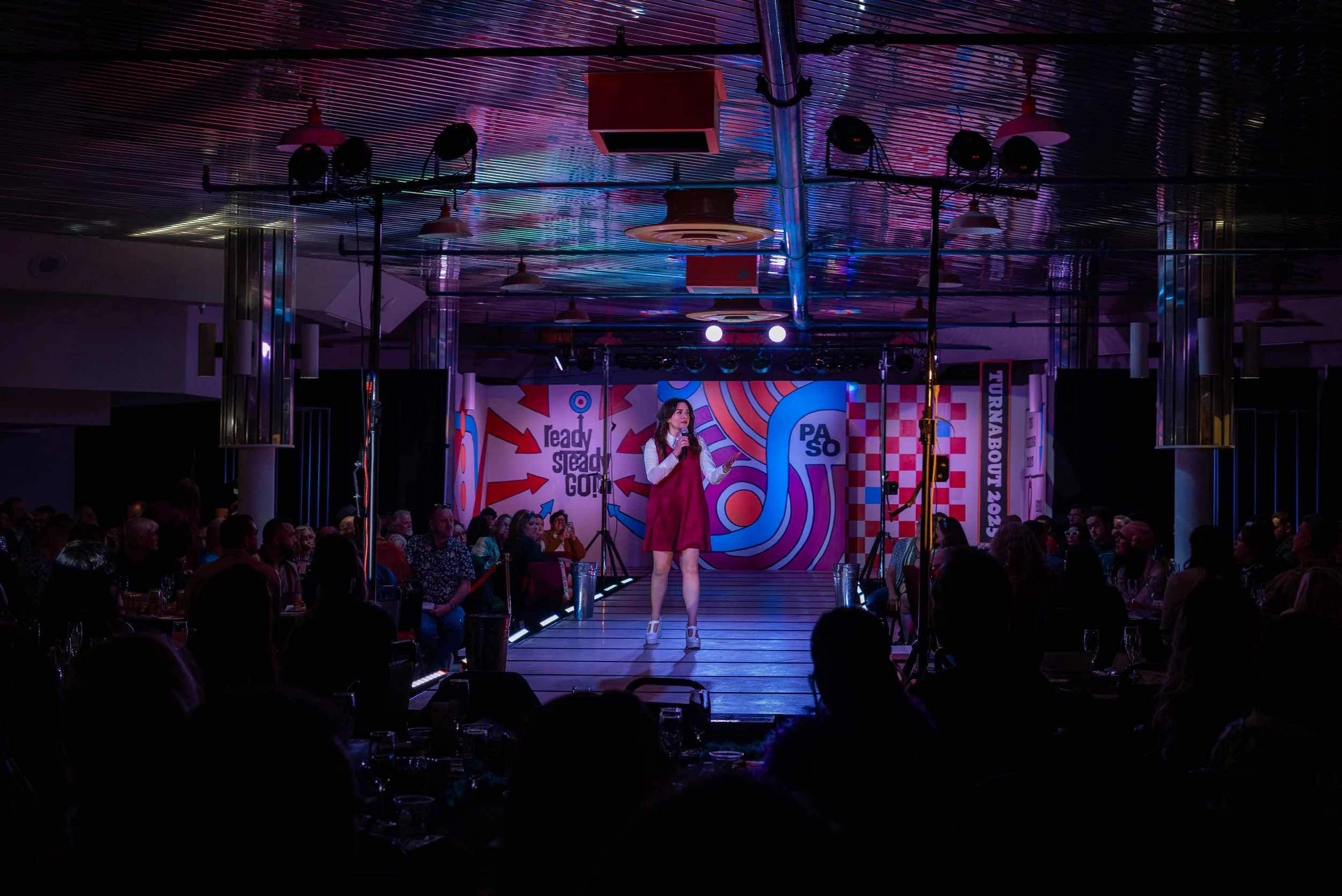 A woman stands on a runway stage with a microphone, performing in front of an audience in a dimly lit venue. The stage has colorful graphic backgrounds with text, and bright lights are directed at her.