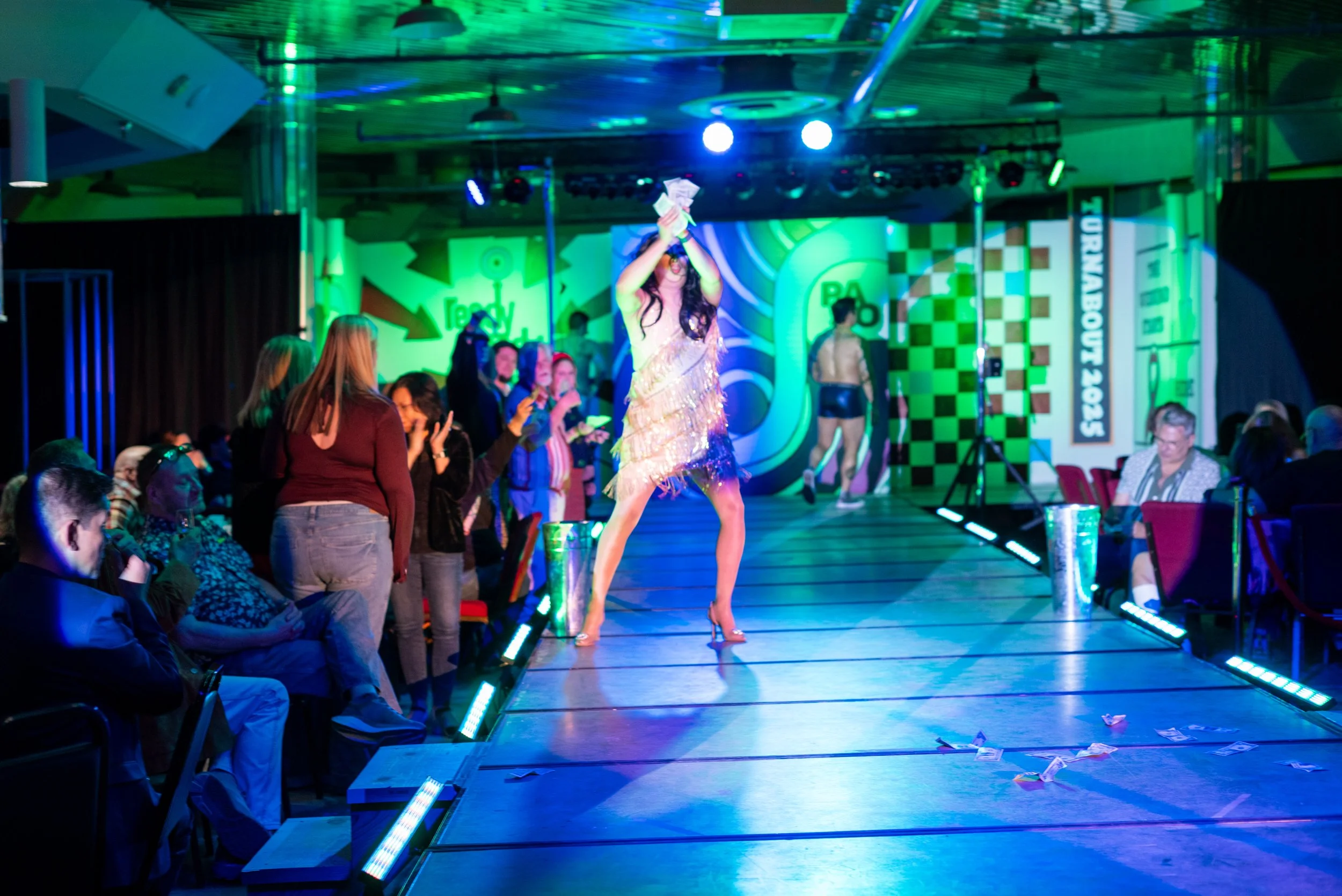 A performer on stage in a glittering dress and high heels holding a stack of money with both hands. The stage has colorful lighting and audiences seated on either side watching, with some people taking pictures or videos.